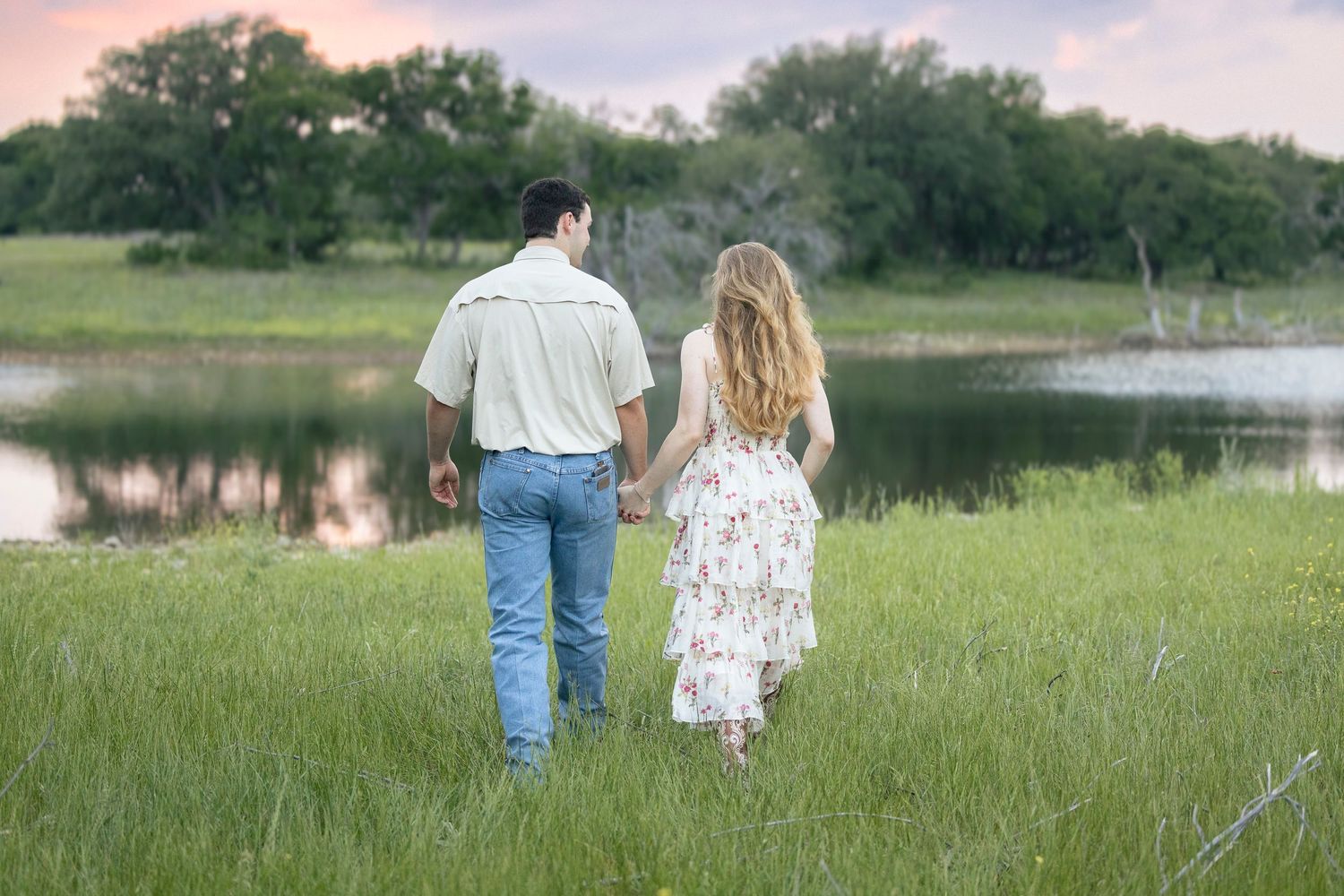 A newly engaged couple walking toward a pink and blue sunset reflecting on a lake.