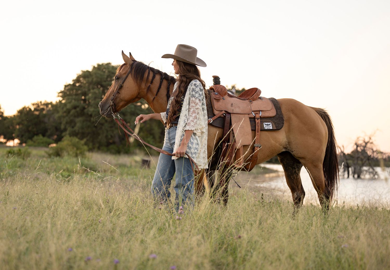 A figure in western attire stands with a brown saddled horse in a grassy field during sunset for senior portraits.