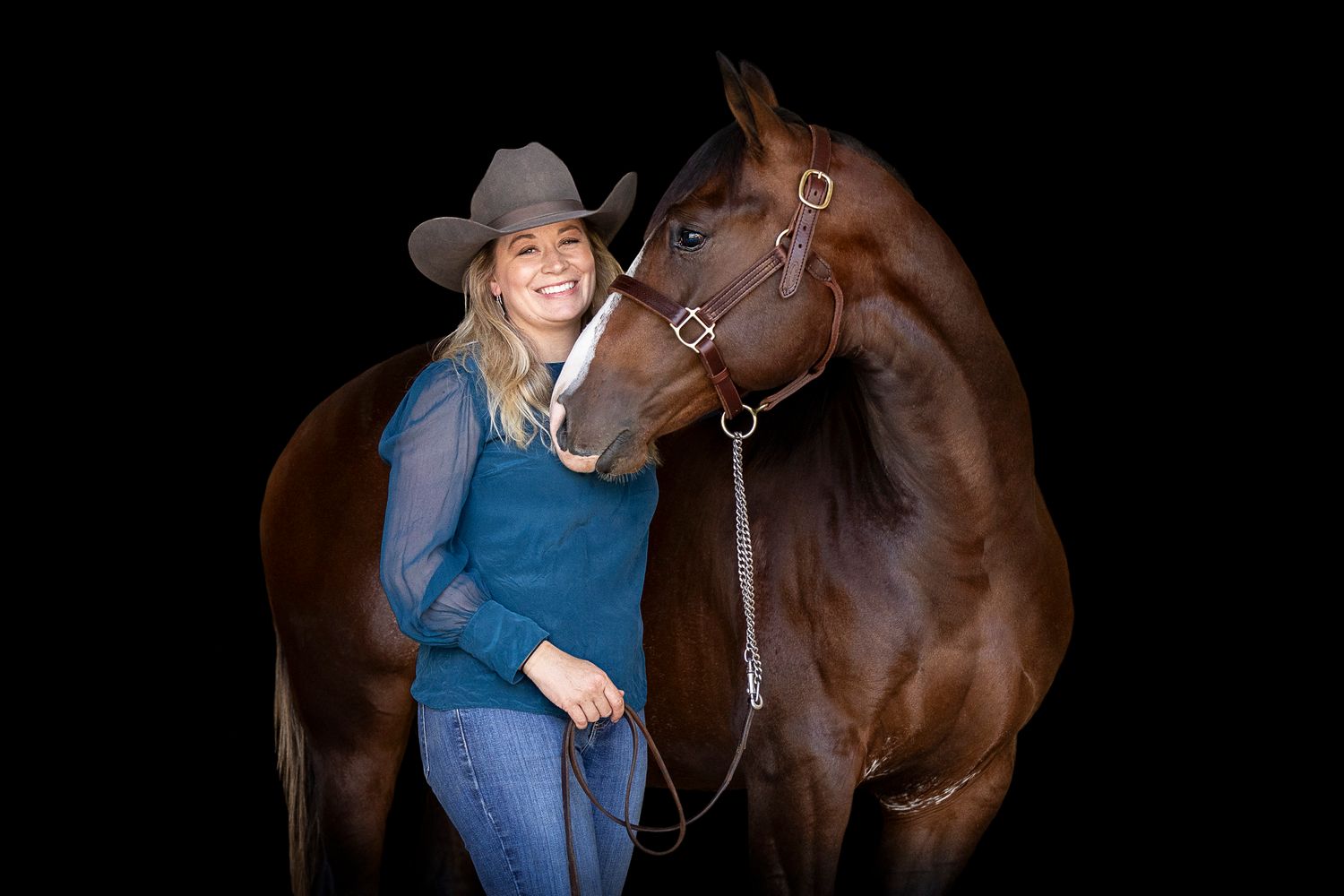 A person wearing a blue shirt and cowboy hat stands with a beautiful dark brown horse against a black background.