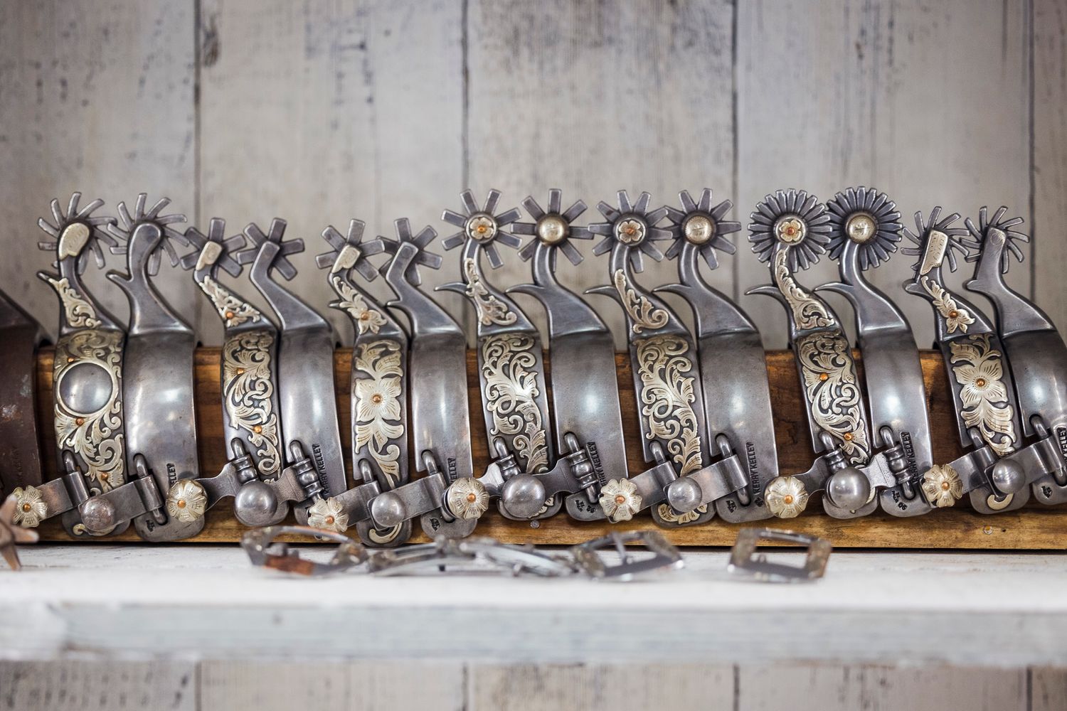 Row of ornate silver spurs with intricate floral designs and metal details displayed on a white shelf.