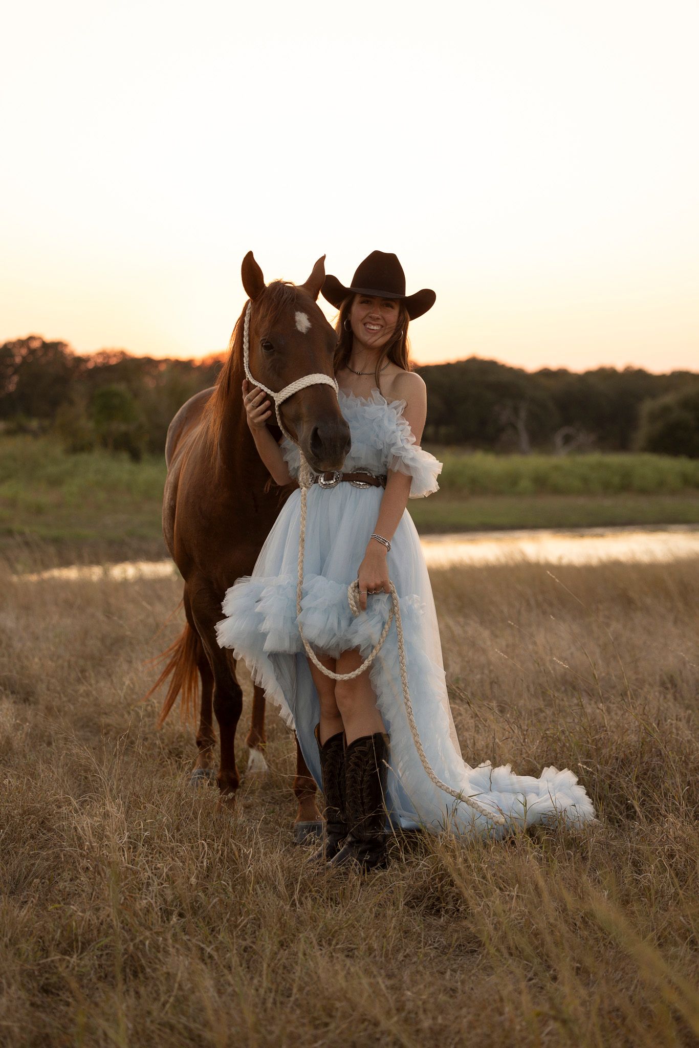 A chestnut horse stands with someone wearing a pale blue dress and cowboy boots in a golden field at sunset.
