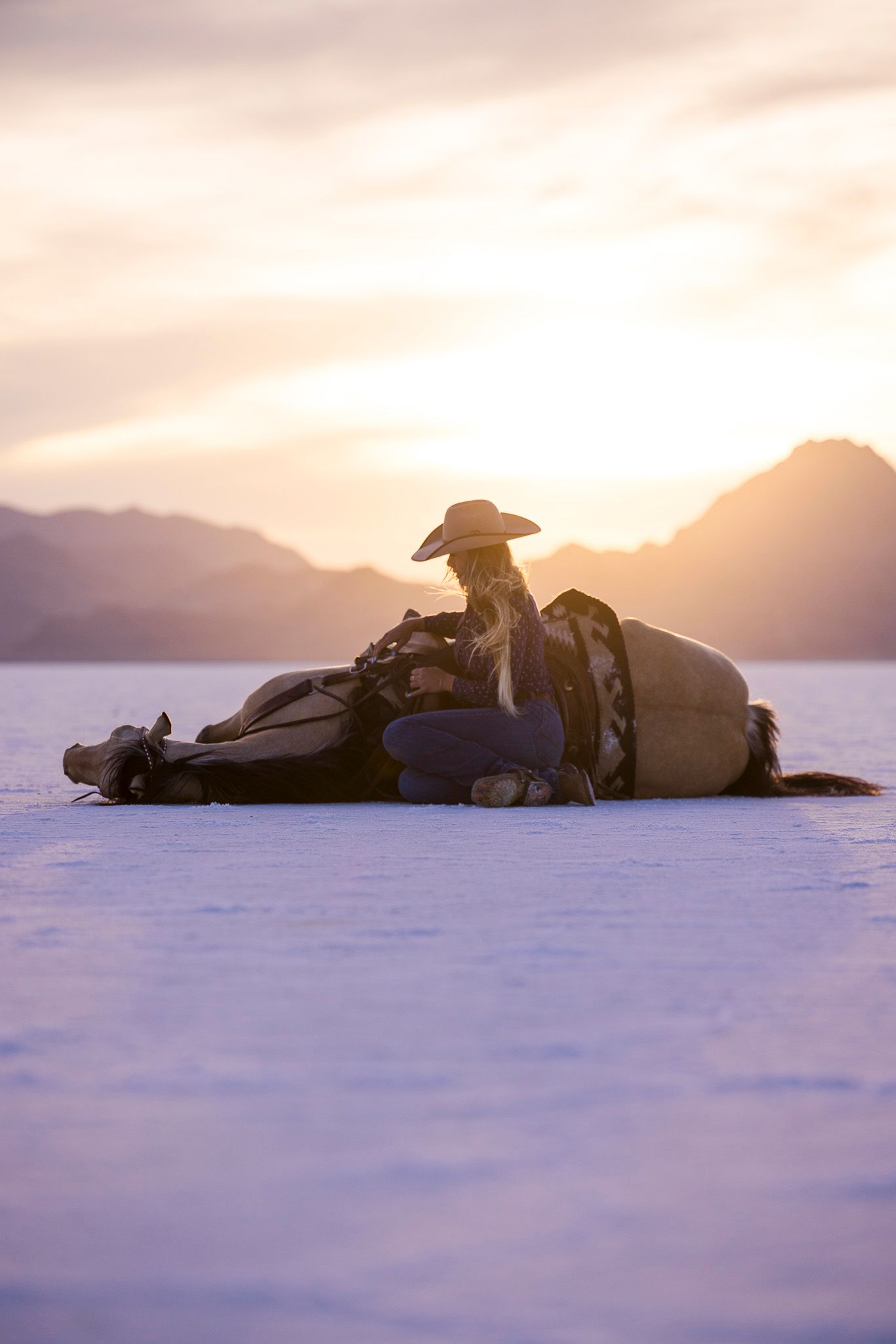 A cowgirl rests against her horse laying on the Salt flats with a purple, orange, and pink sunset.
