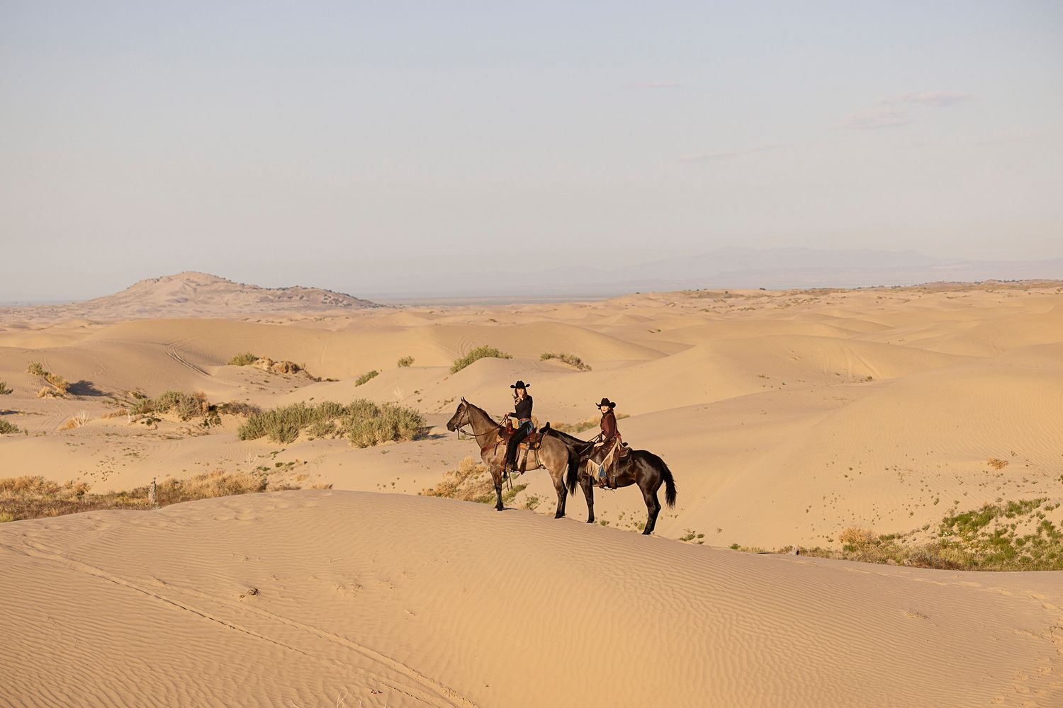 Two people on horseback traverse golden sand dunes in a vast desert landscape during golden hour.