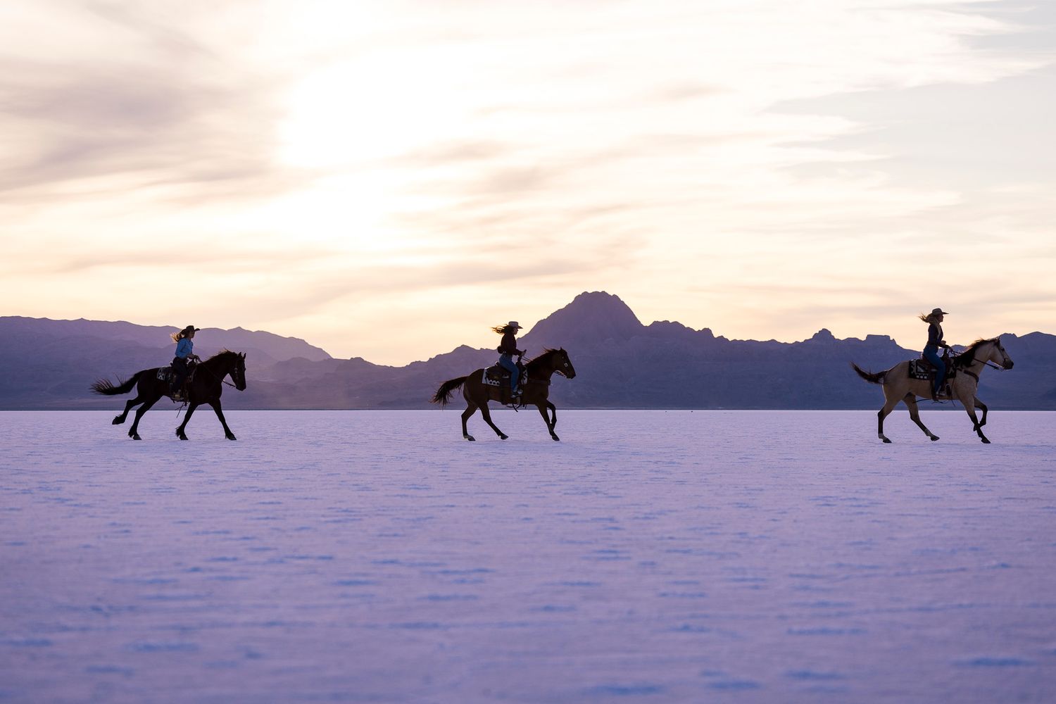 Three horse riders gallop across the Salt Flats against a backdrop of mountains and sunset sky in silhouette.