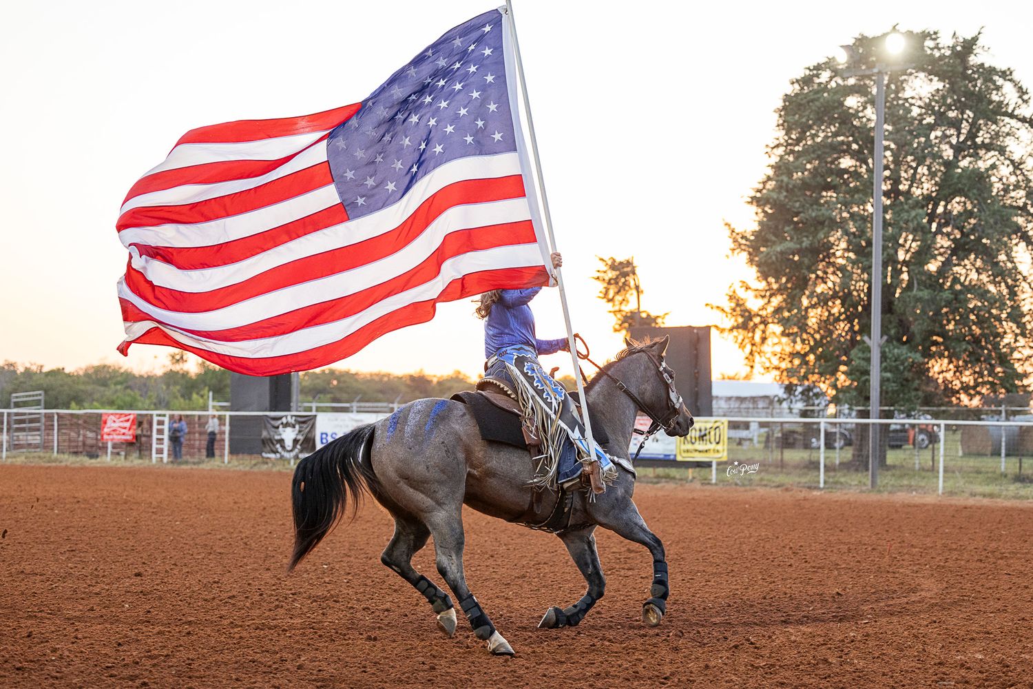 A rider carries the American flag while galloping on horseback across a dirt arena at sunset in Rockdale, Texas