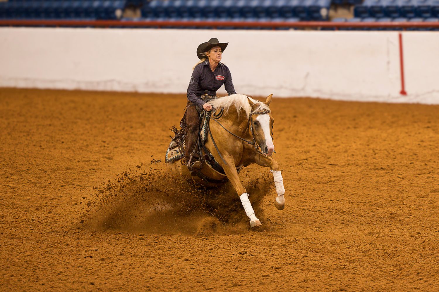 Horse and rider perform sliding stop maneuver in the John Justin Arena kicking up dirt cloud on reddish ground.