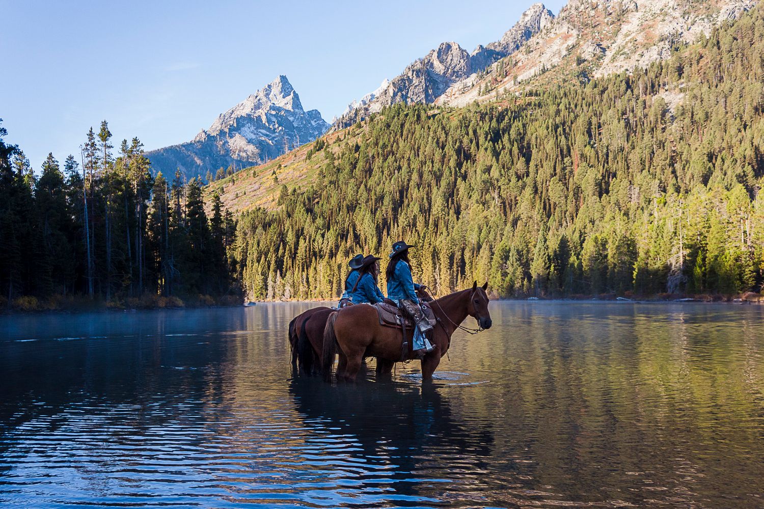 3 horseback riders wade through a serene mountain lake surrounded by evergreen forest and rocky peaks at sunset.