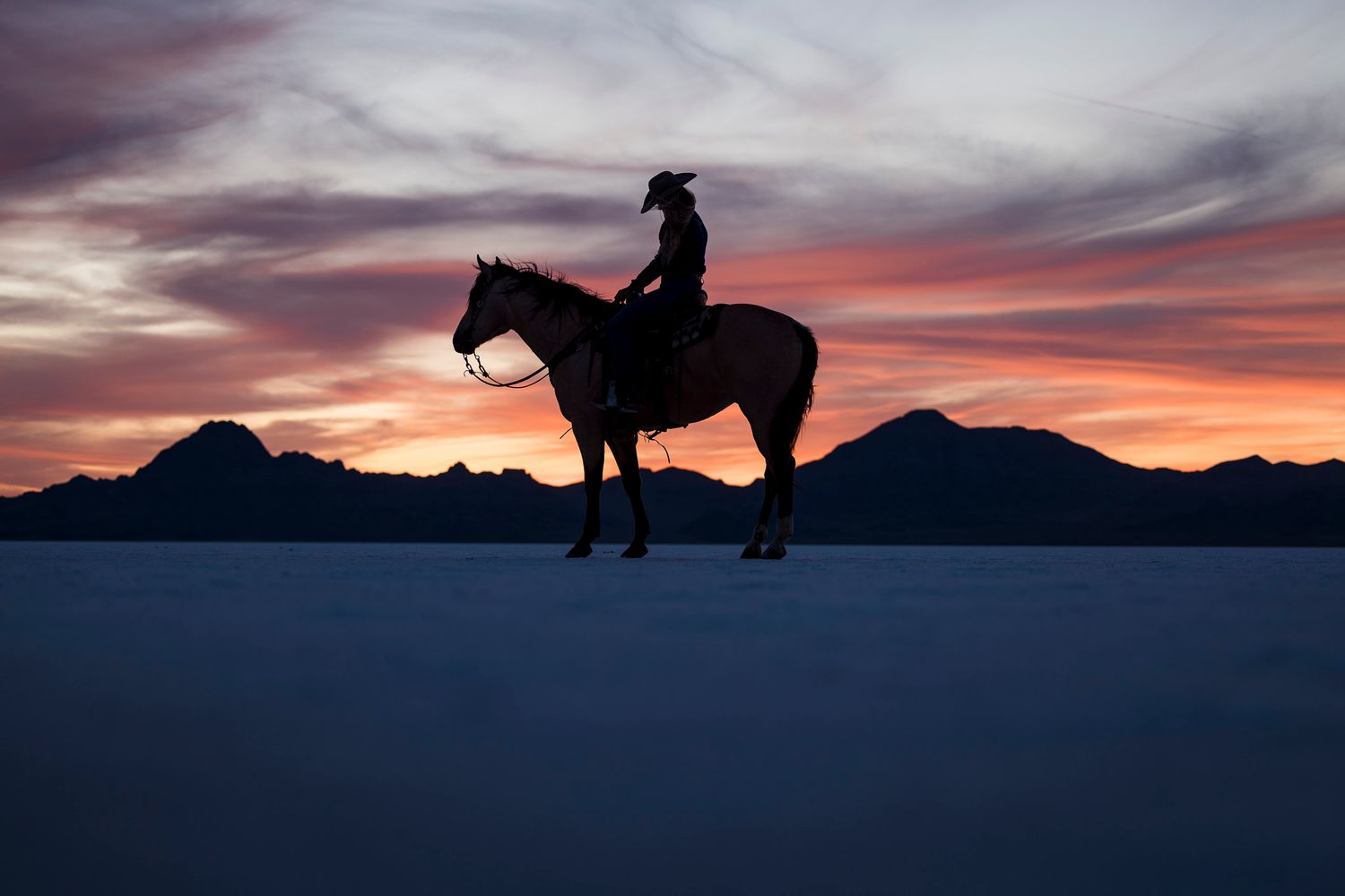 Silhouette of a horseback rider against a dramatic sunset sky with mountain backdrop on the Bonneville Salt Flats.