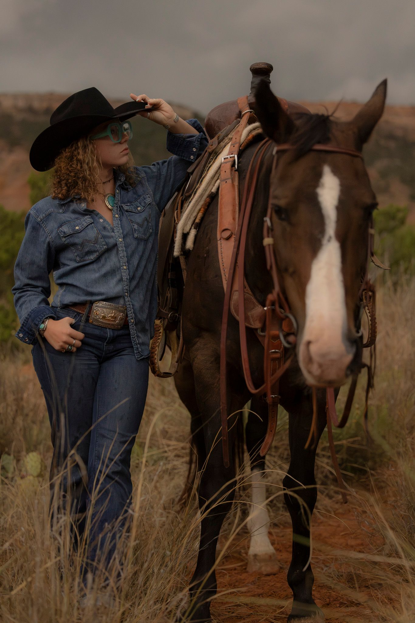 A cowgirl wearing denim and turquoise posed with a bay horse as a thunderstorm is rolling into the Palo Duro Canyon State Park