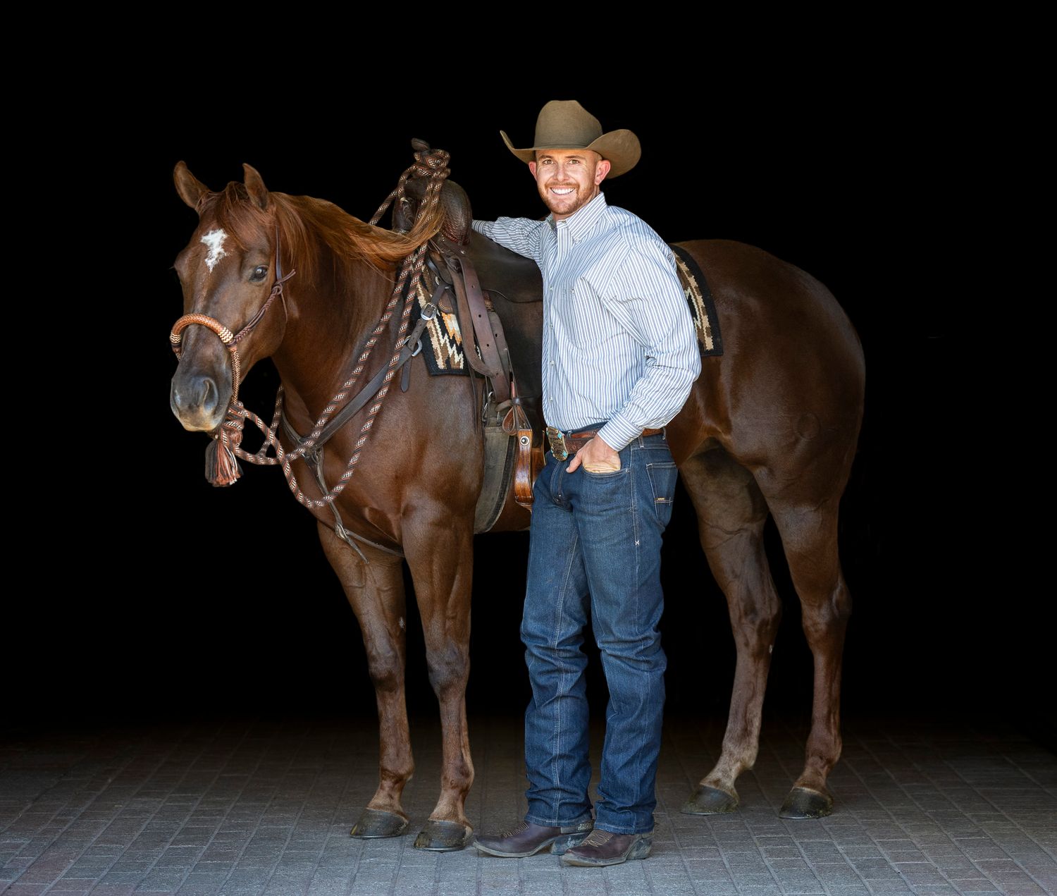 A black background portrait of a trainer and their sorrel horse. The horse is saddled and wearing a Hackamore