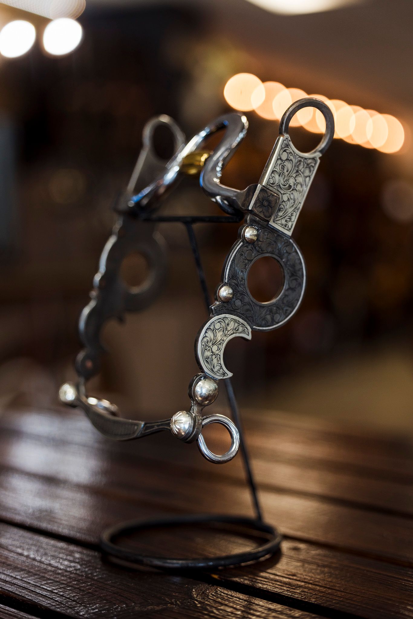 Close-up of a decorative horse bit with silver accents and ornate details displayed on a wooden stand with bokeh lights.