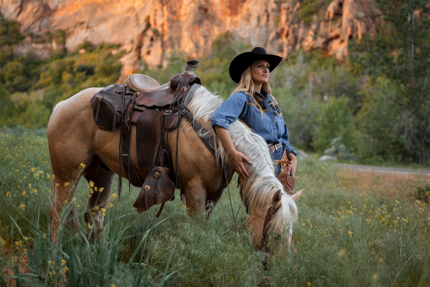 Western rider in denim and cowboy hat rests with saddled horse grazing in meadow against red rock cliffs at sunset.