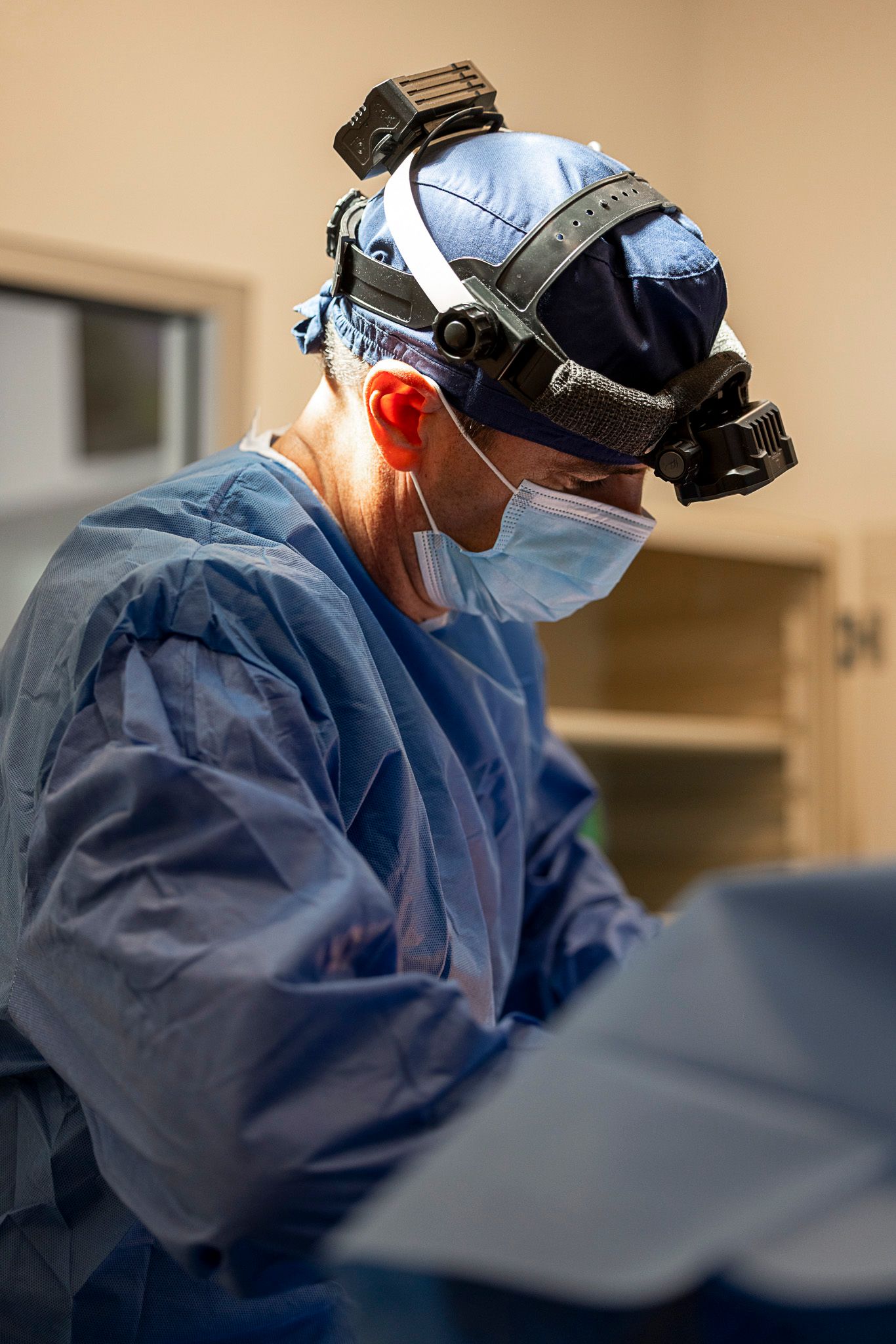 Veterinary surgeon in blue scrubs and surgical mask performs medical procedure while wearing head-mounted equipment.