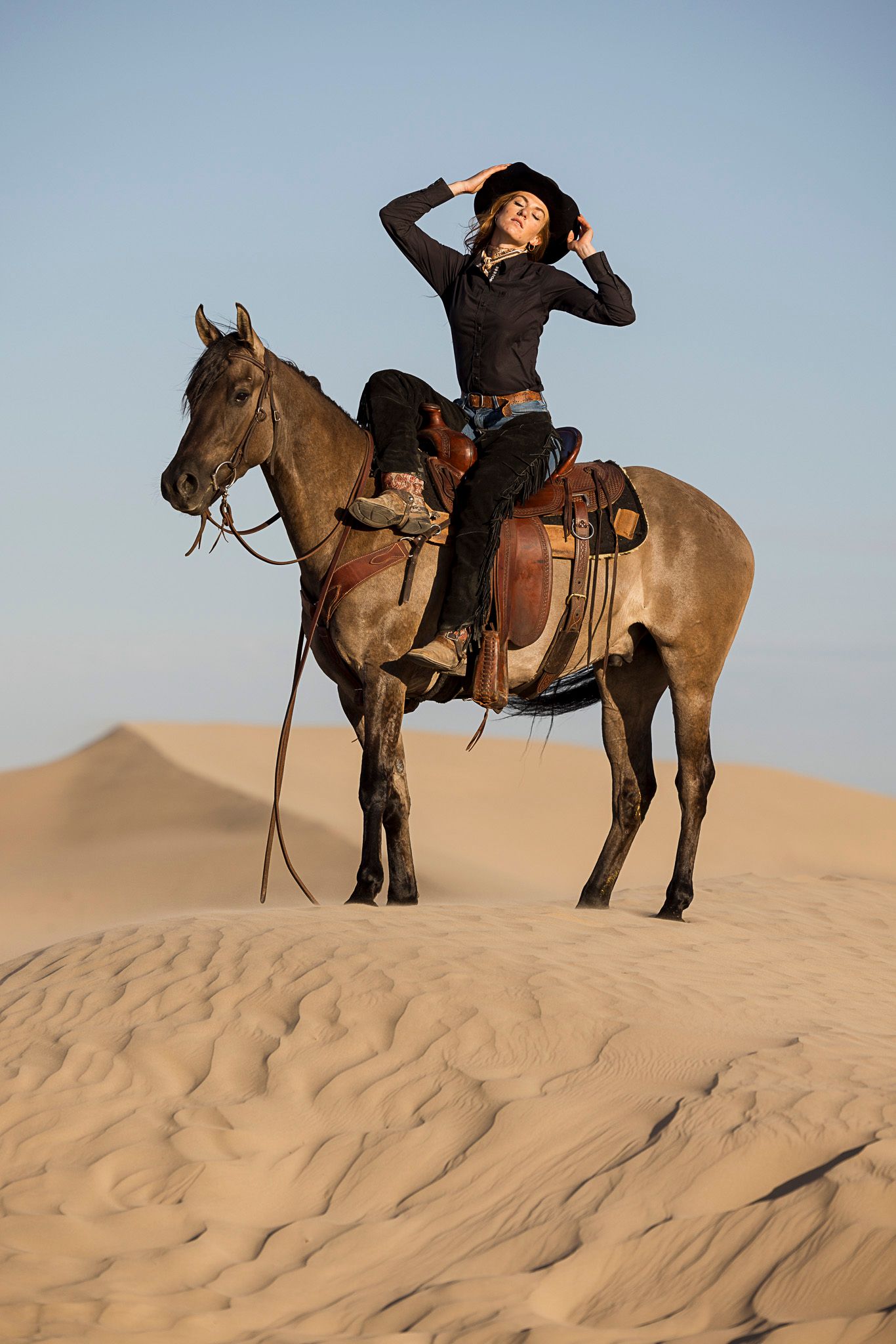 A rider in western attire poses atop a horse on a sand dune in the desert during golden hour.