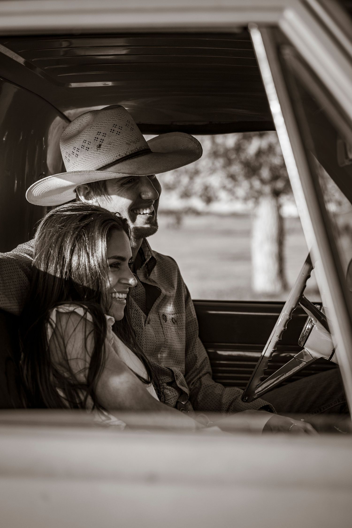 Black and white photo showing someone wearing a cowboy hat behind the steering wheel of a vintage vehicle.