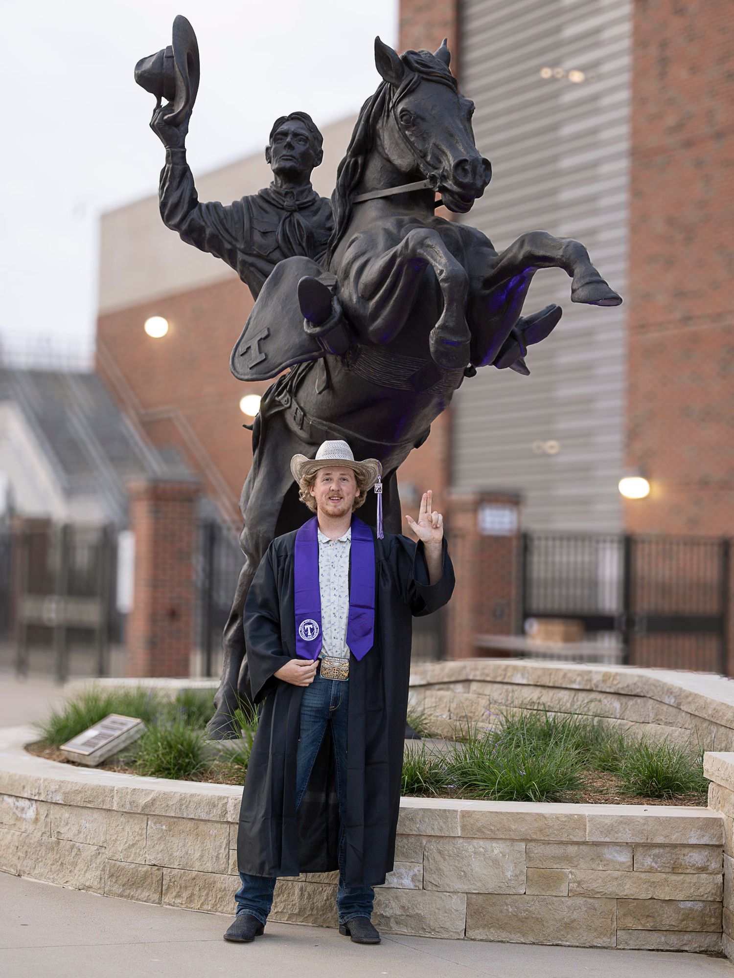 A graduate stands in front of a dramatic bronze equestrian statue on a stone pedestal wearing academic regalia with Tarleton purple sash.