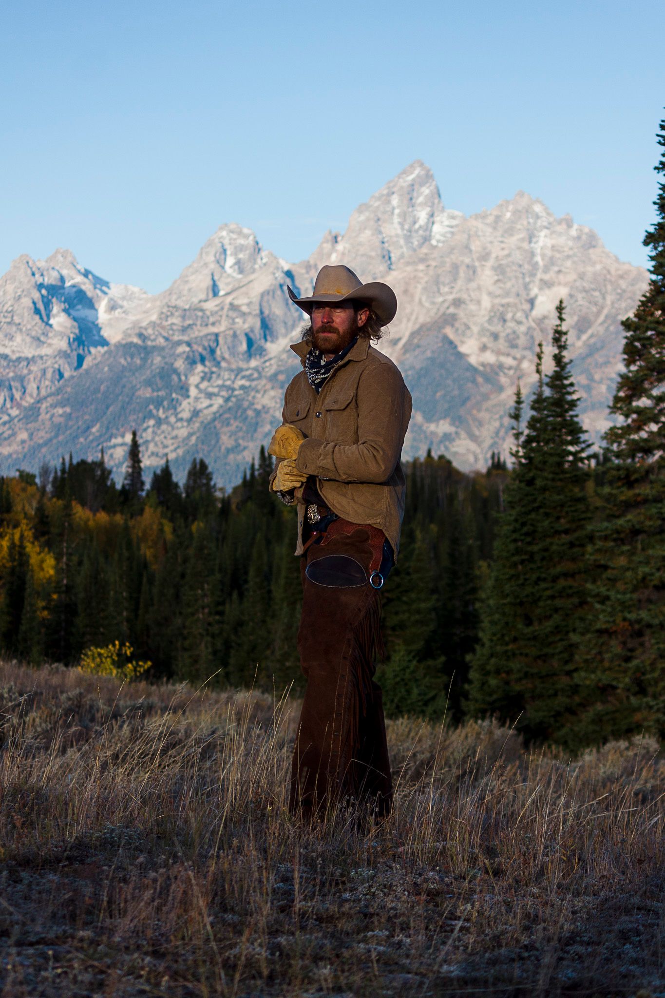 A Yellowstone cowboy wearing western attire standing in a field with the majestic Grand Teton mountains rising in the background.