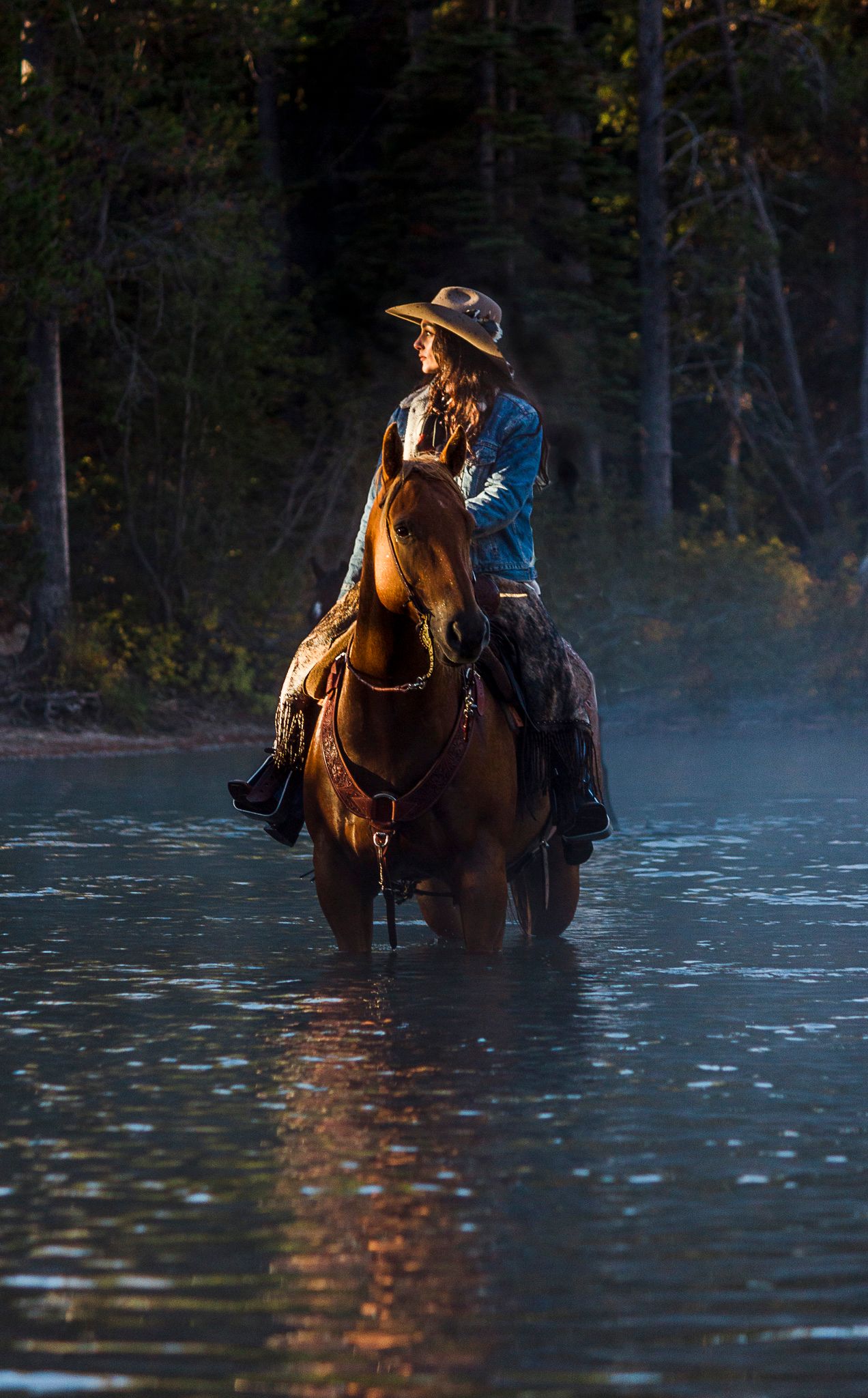 A cowgirl wearing a hat riding horseback through misty water in early morning light in Grand Teton National Park