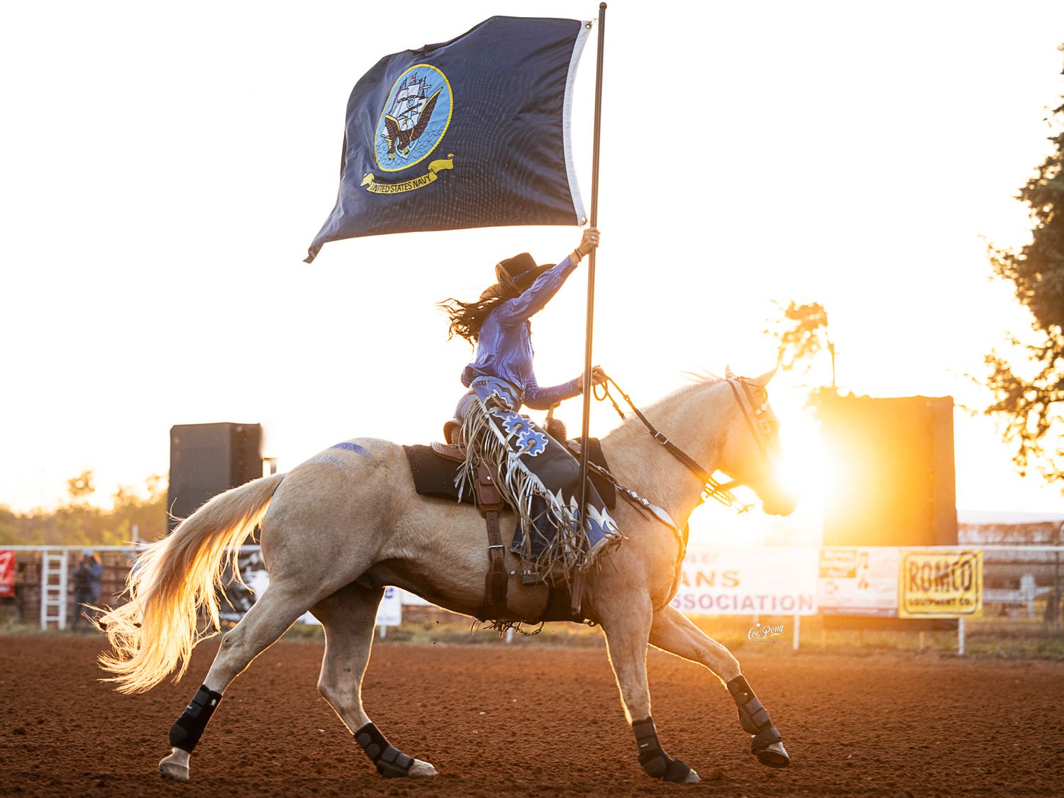 Drill Team rodeo rider gallops on horseback carrying Navy flag during sunset in an arena.