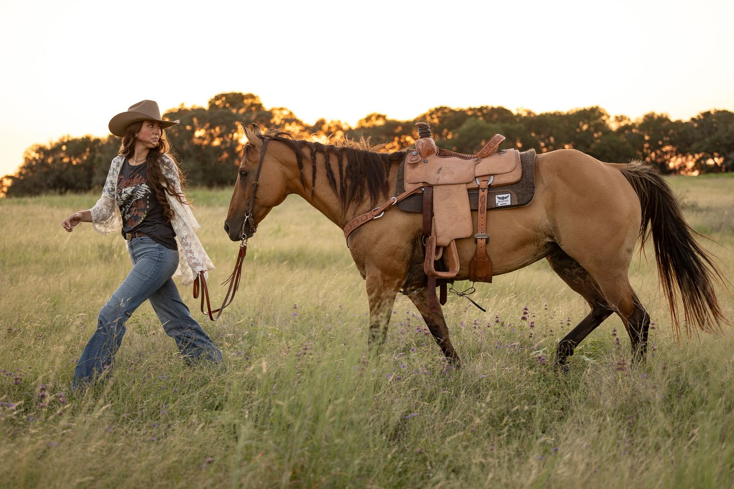 A western figure walks through a grassy field at sunset leading a saddled brown horse.