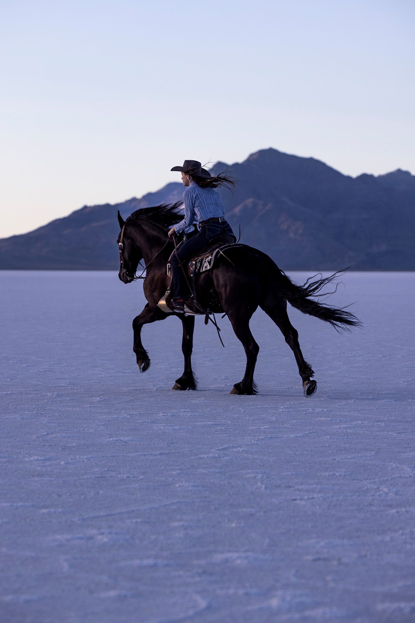 A rider on horseback gallops across a stark salt flat with mountain silhouettes in the twilight background.