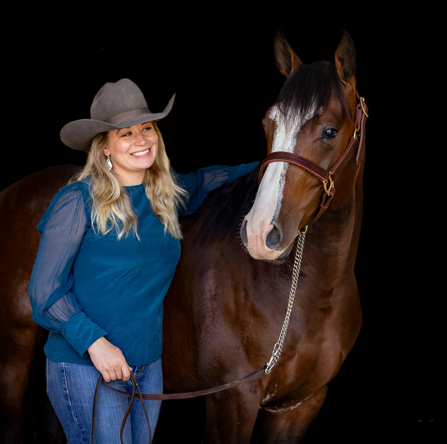 A bay horse with white blaze stands next to someone in a cowboy hat and blue top in a dark studio setting.