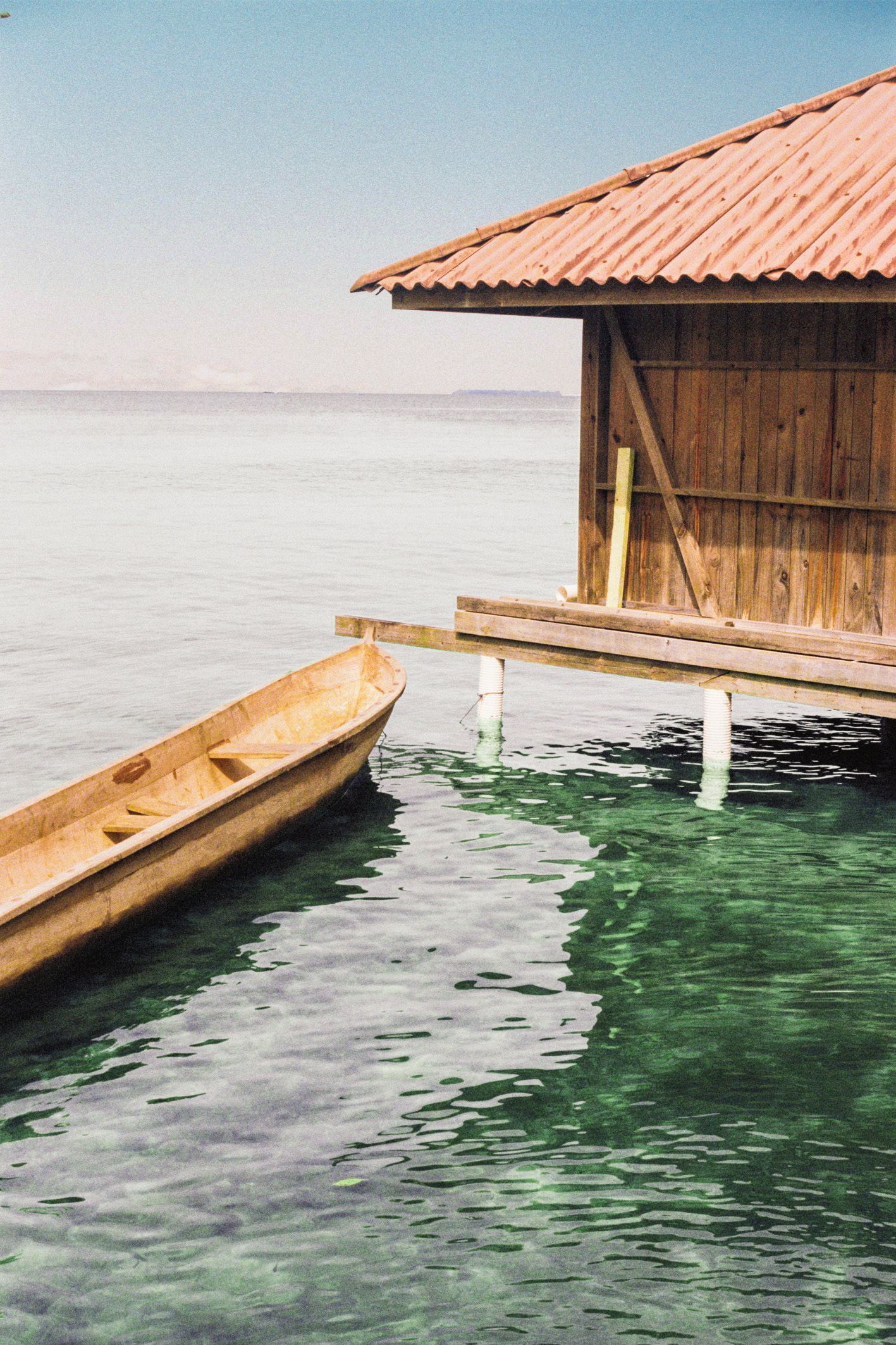 A wooden boat floats on calm, teal-colored water near a small, wooden, elevated structure. 35mm film shot taken in Bocas del Toro, Panama