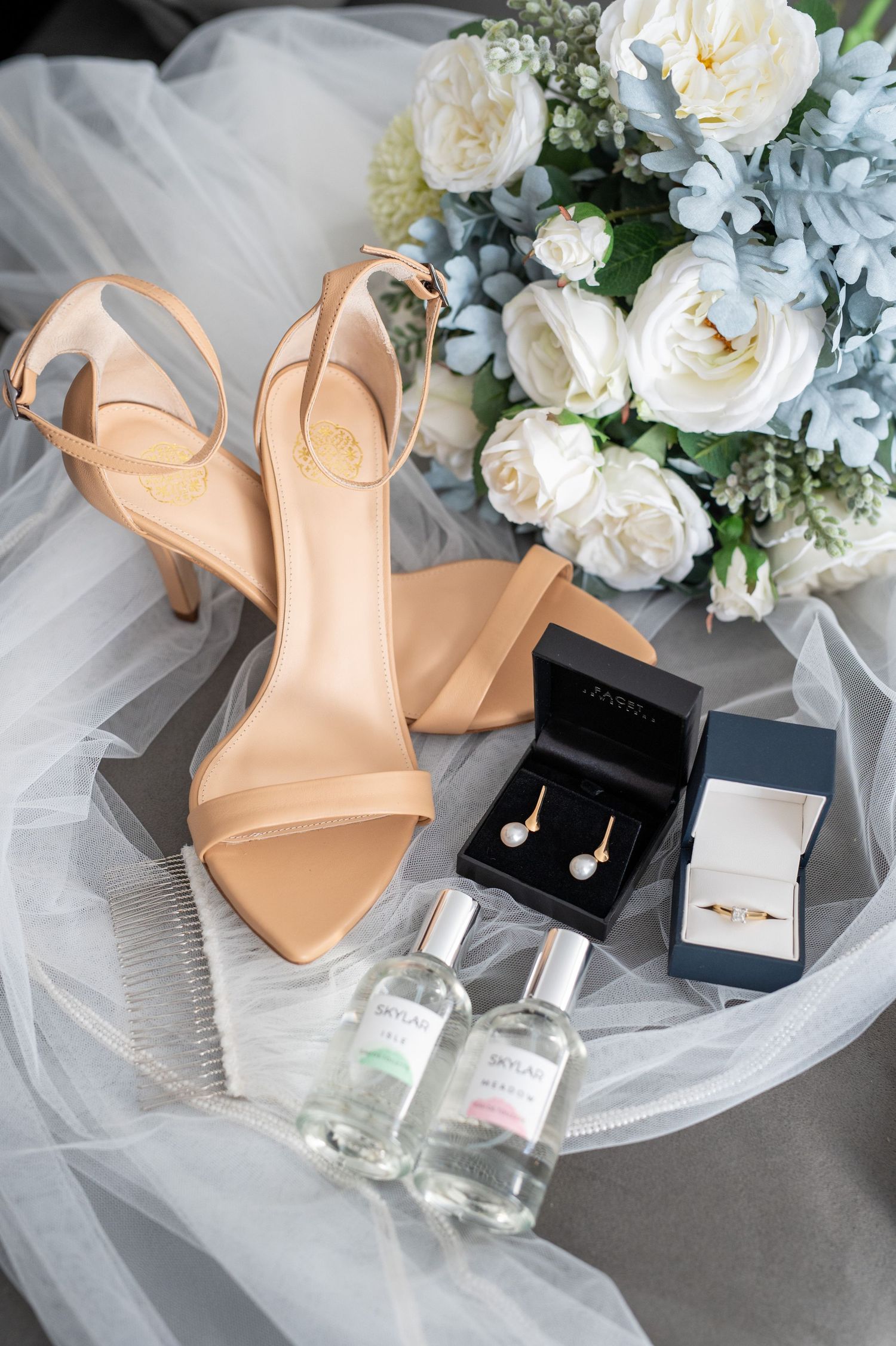 Wedding details featuring nude heels, jewelry boxes, perfume bottles and a white and blue floral bouquet on tulle fabric.