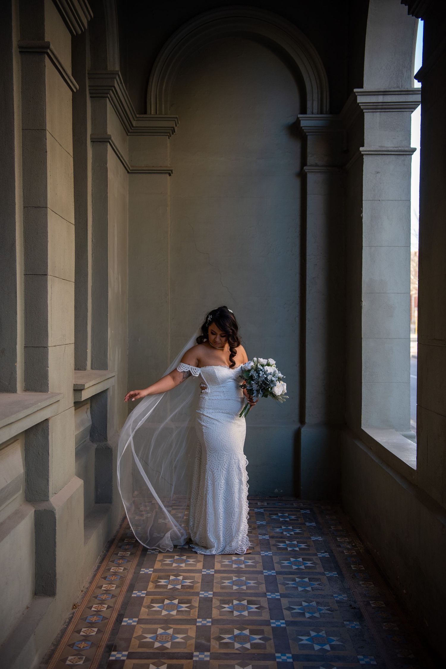 A bride in a flowing white dress stands gracefully in an elegant arched hallway with patterned tile flooring.
