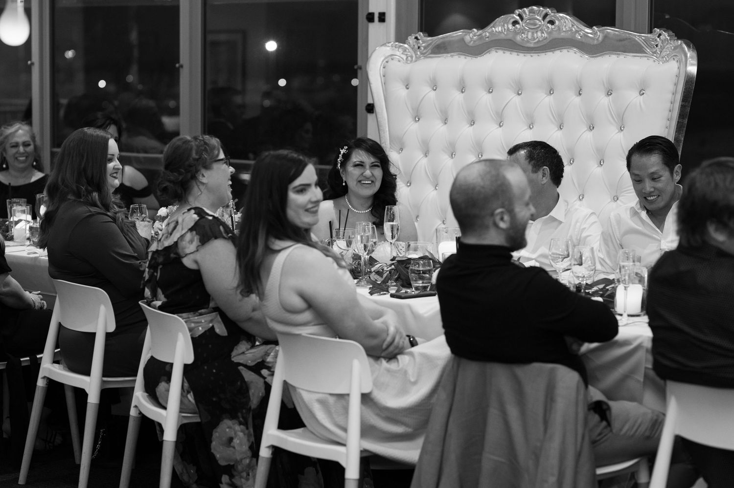 Wedding guests enjoying dinner and conversation at an elegant reception with a white tufted throne chair visible in black and white.