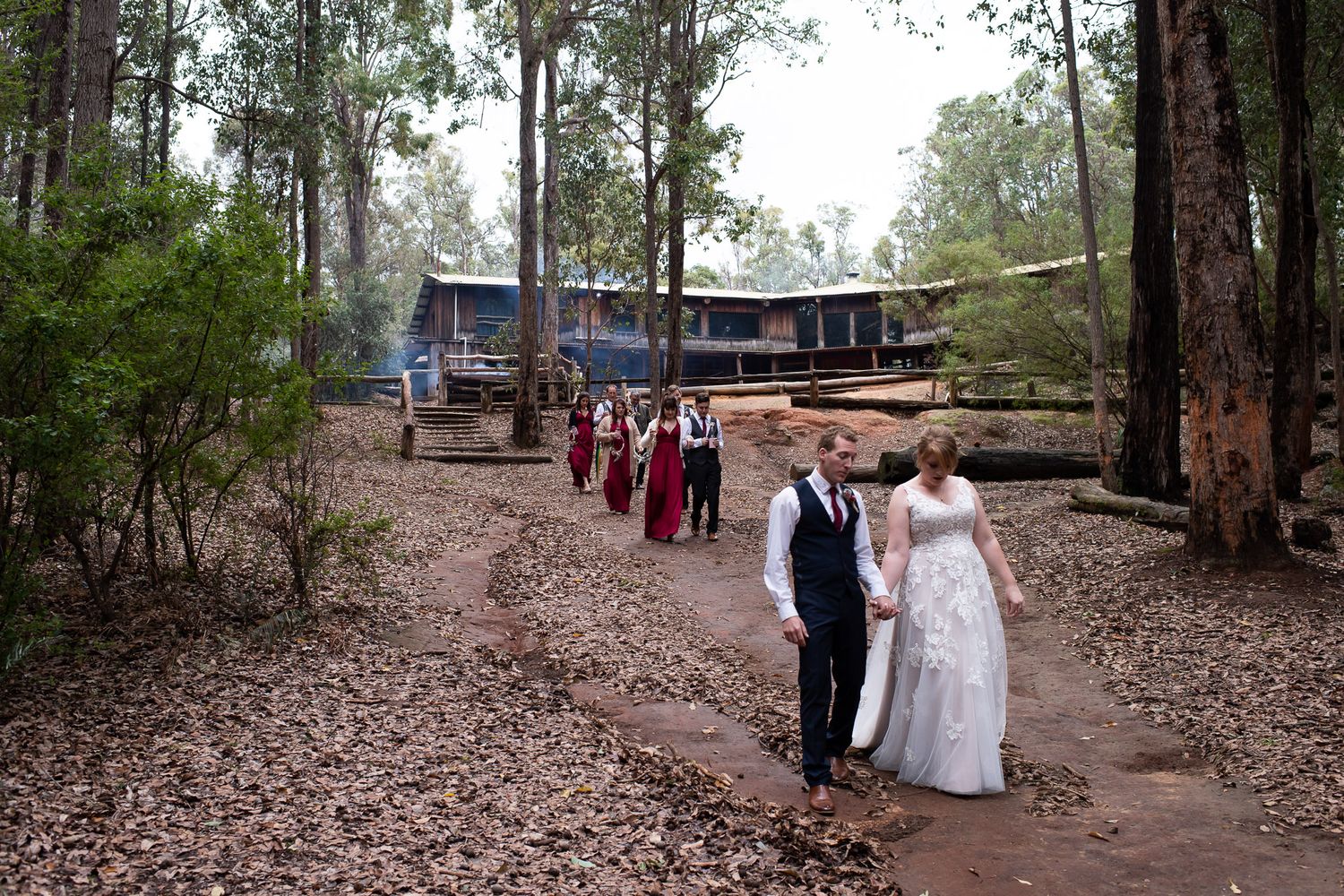 A couple in formal wedding attire walks along a dirt path through a wooded area with rustic buildings in the background.