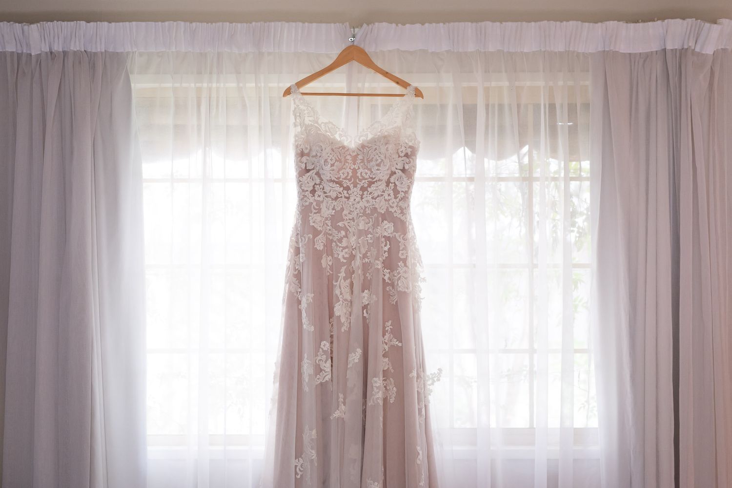 A delicate blush wedding dress with lace details hangs on a wooden hanger against sheer white curtains in natural light.