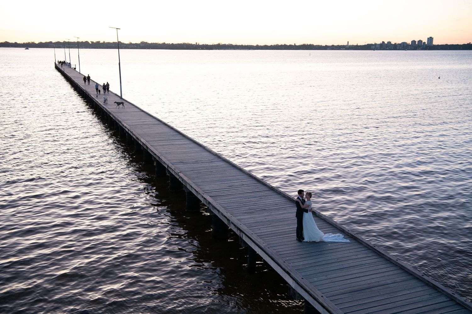 Long wooden pier extends over calm water at sunset with distant figures.