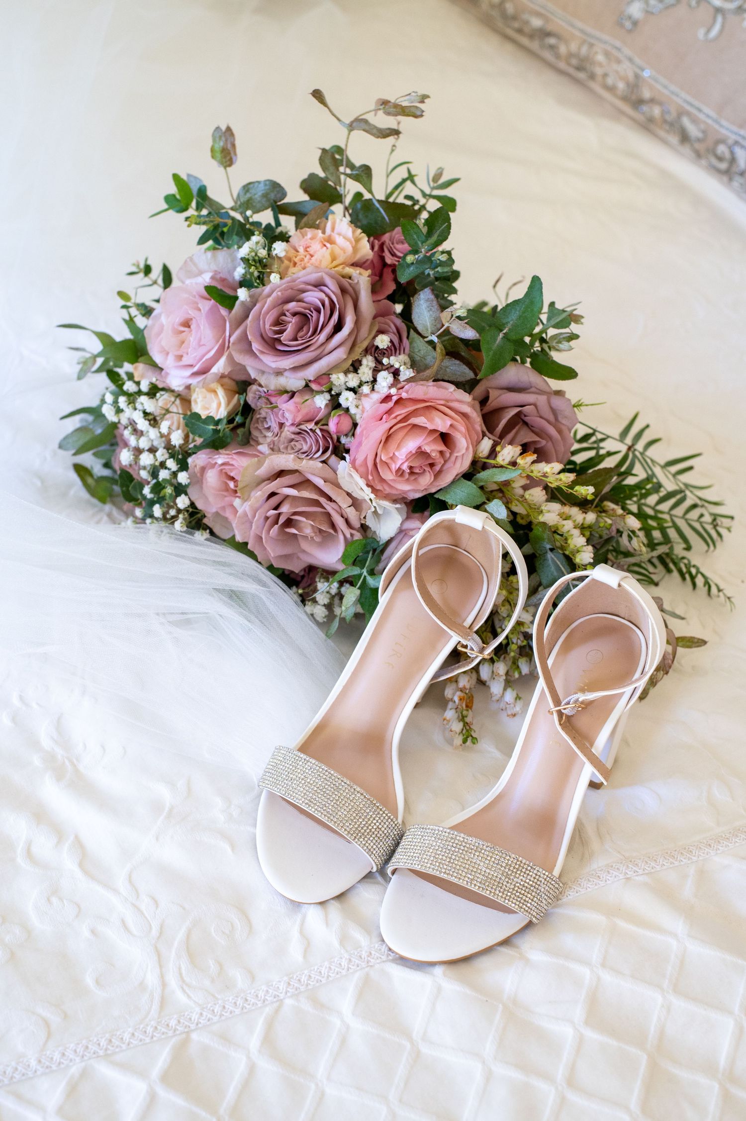 Elegant bridal bouquet of pink and purple roses with white heels featuring sparkling embellishments on white bedding.