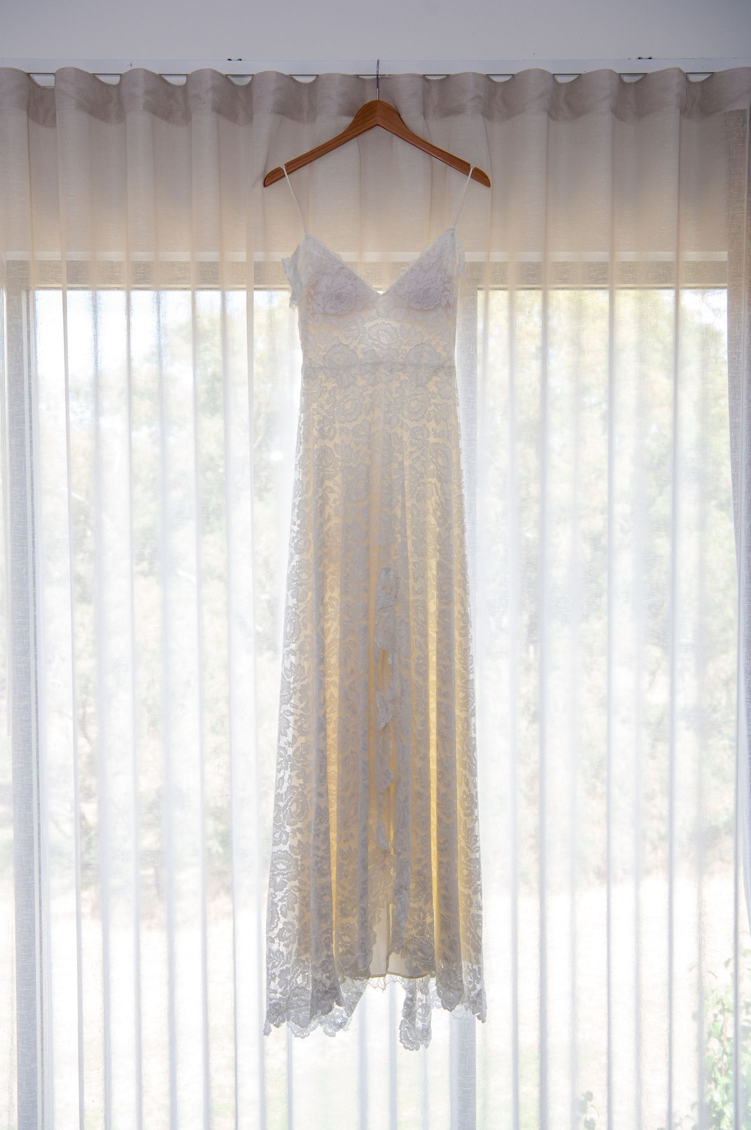 White lace wedding dresses hanging on wooden hangers against sheer curtains in a bridal suite.