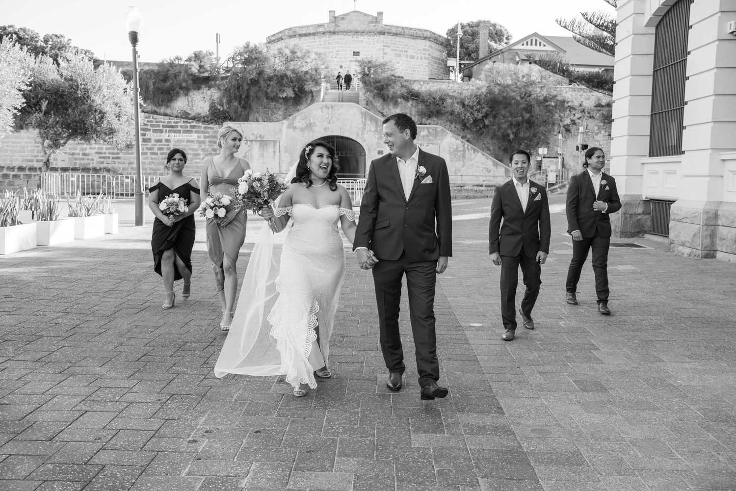 A wedding party walks together along a stone pathway outside the Roundhouse in Fremantle, Perth on a sunny day.