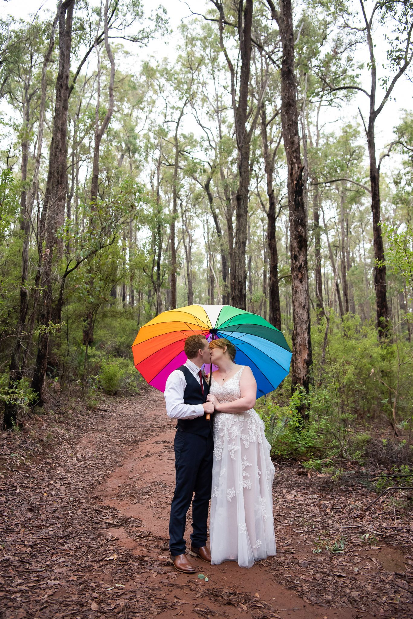A couple shares a romantic moment under colorful umbrellas on a wooded trail.