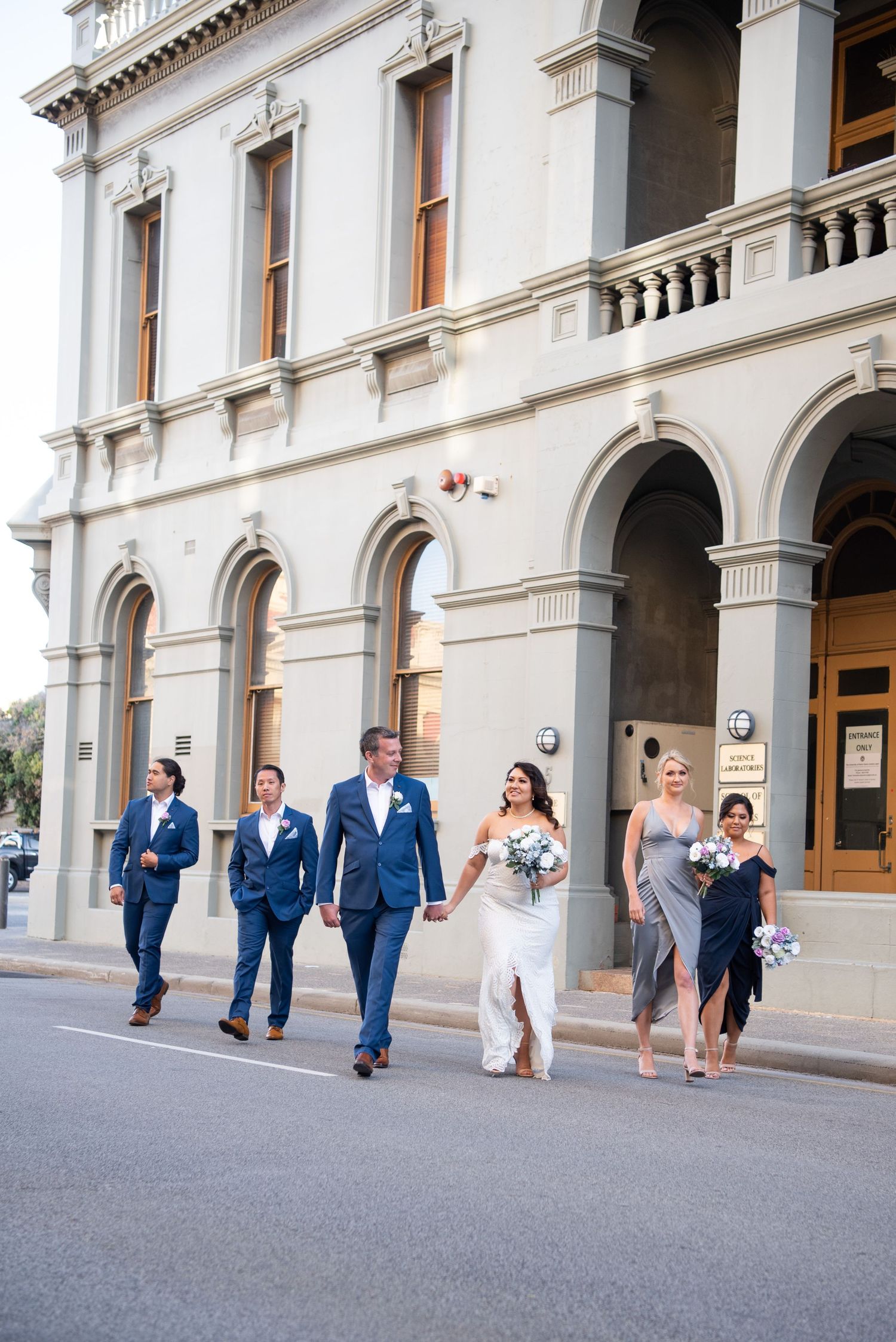 Wedding party walks along vintage building exterior with classical architecture.
