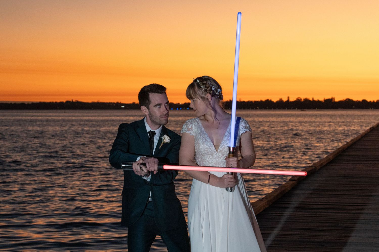 Wedding couple silhouetted against dramatic orange sunset over waterfront dock.