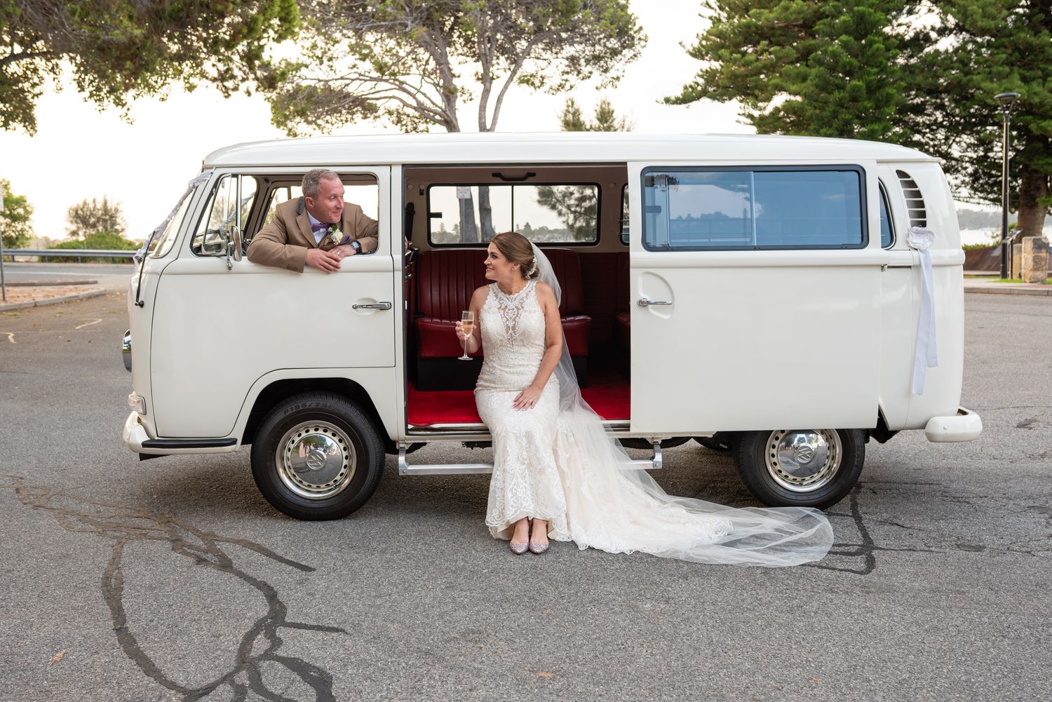 Fun wedding portrait of groom looking out of passenger window of white Kombi at his bride who is sitting in the open door sipping champagne