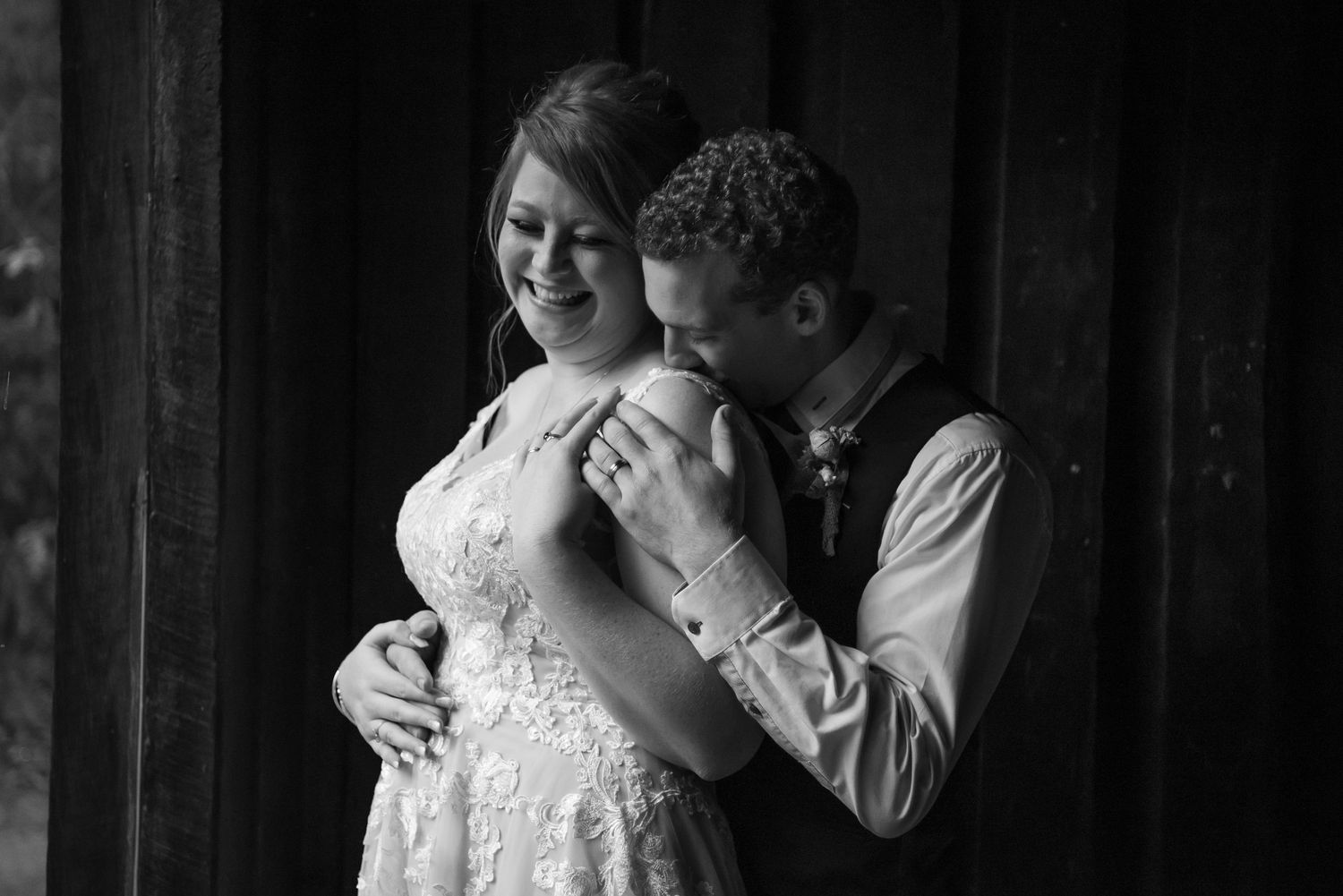 Intimate black and white wedding portrait shot of couple embracing against rustic barn wall.