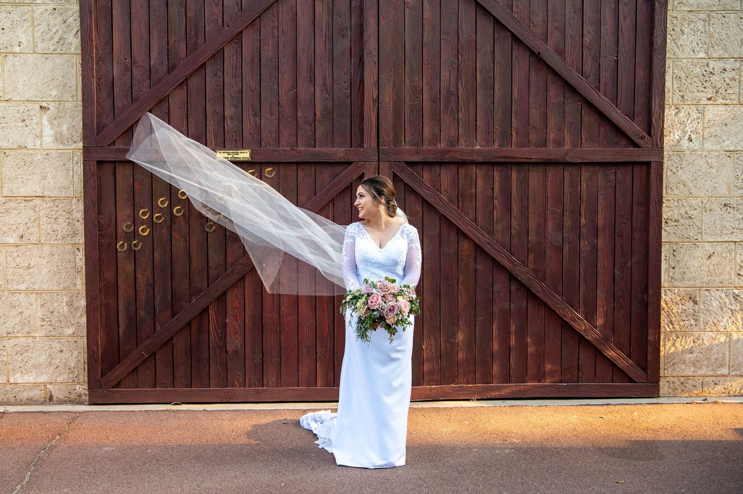 A bride smiles looking at her veil blowing away from her in the wind standing infront of a rustic wooden barn door