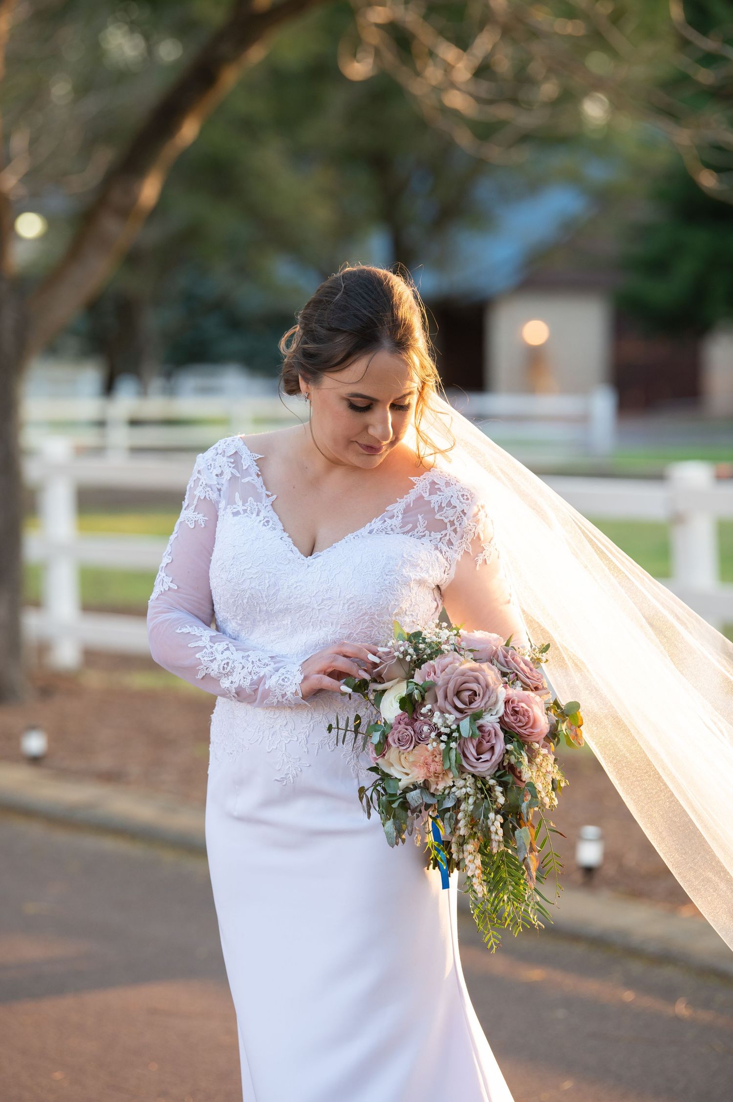 Bride in long-sleeved lace wedding gown holds cascading bouquet of pink and mauve roses during sunset outdoor session.