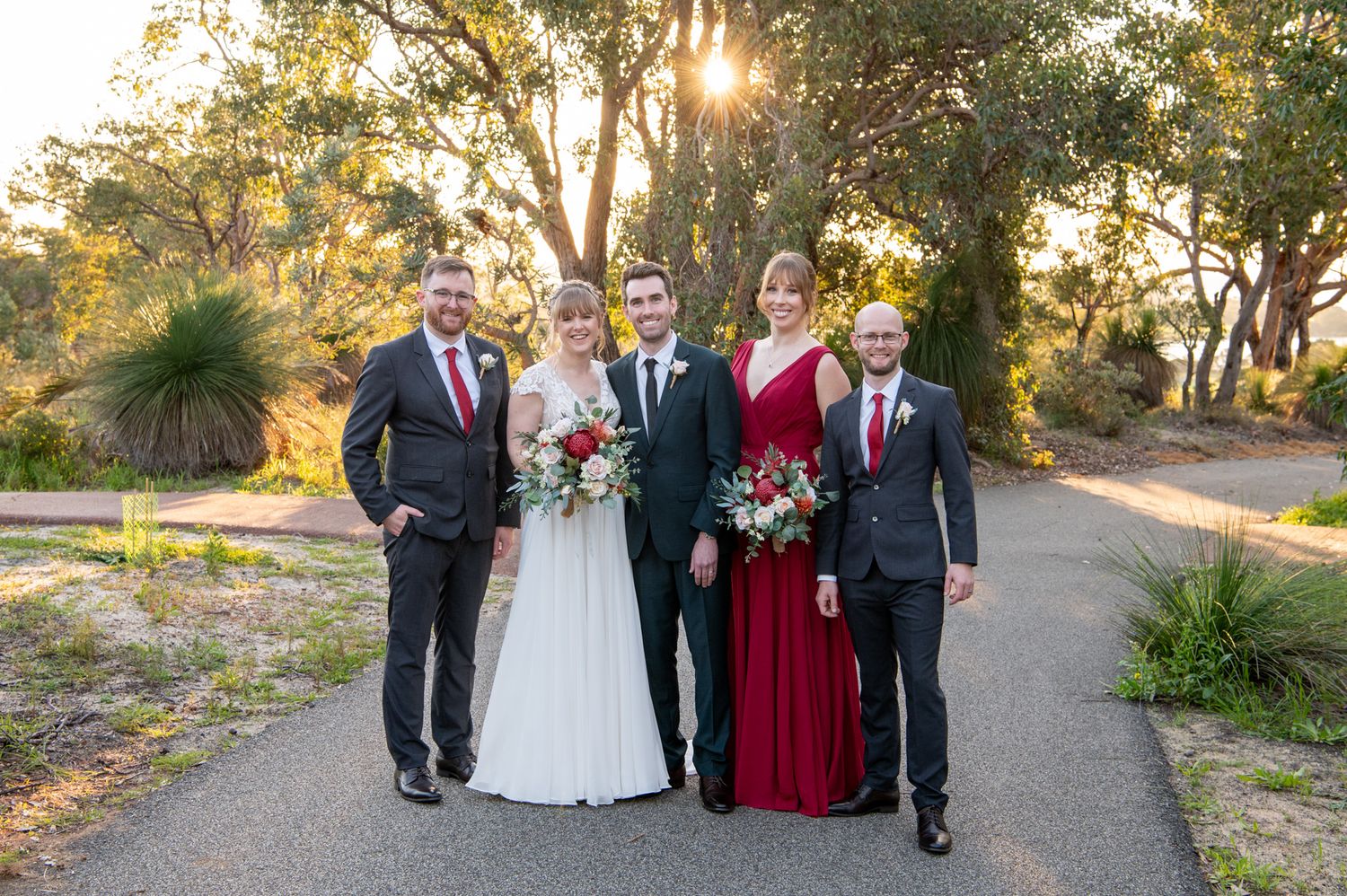 Wedding party stands together on a sunlit path with trees in background during golden hour sunset.
