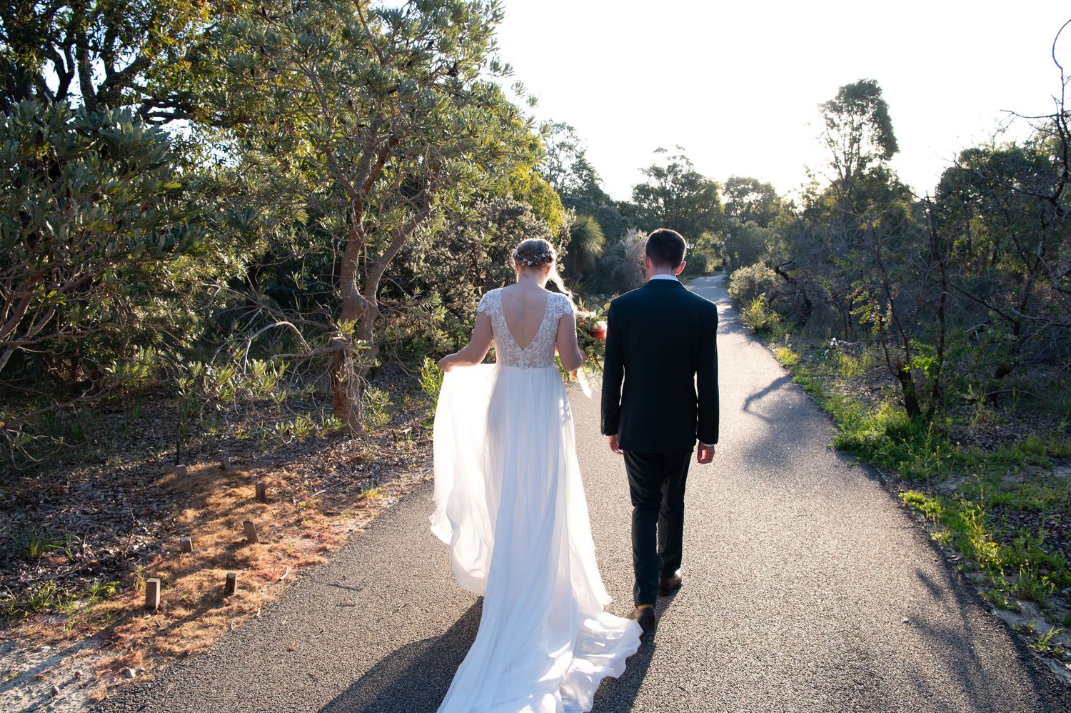 Newly married couple walks hand in hand along a scenic path surrounded by greenery.