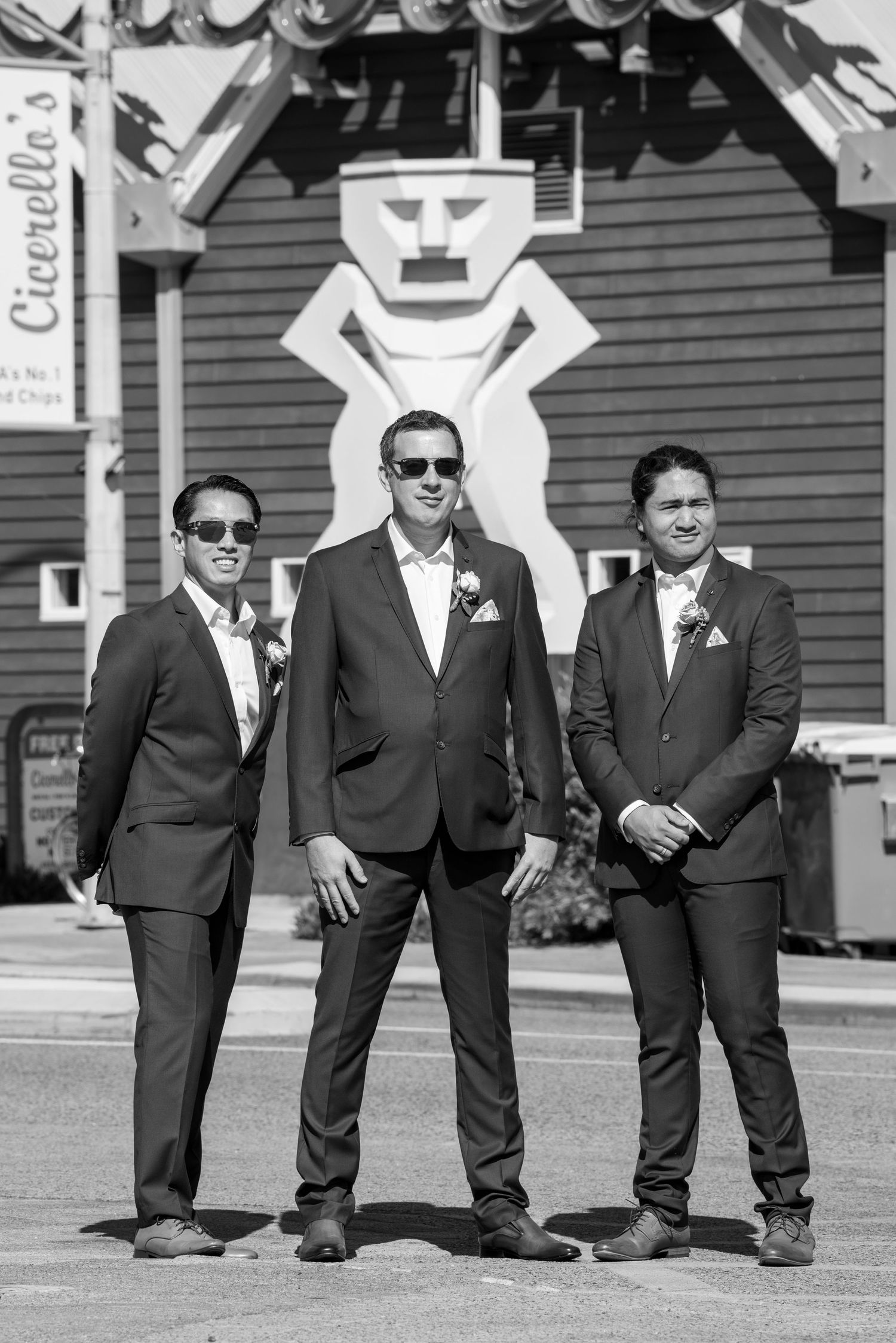 Groomsmen in dark suits and sunglasses lined up in casual pose outdoors in black and white wedding photograph.