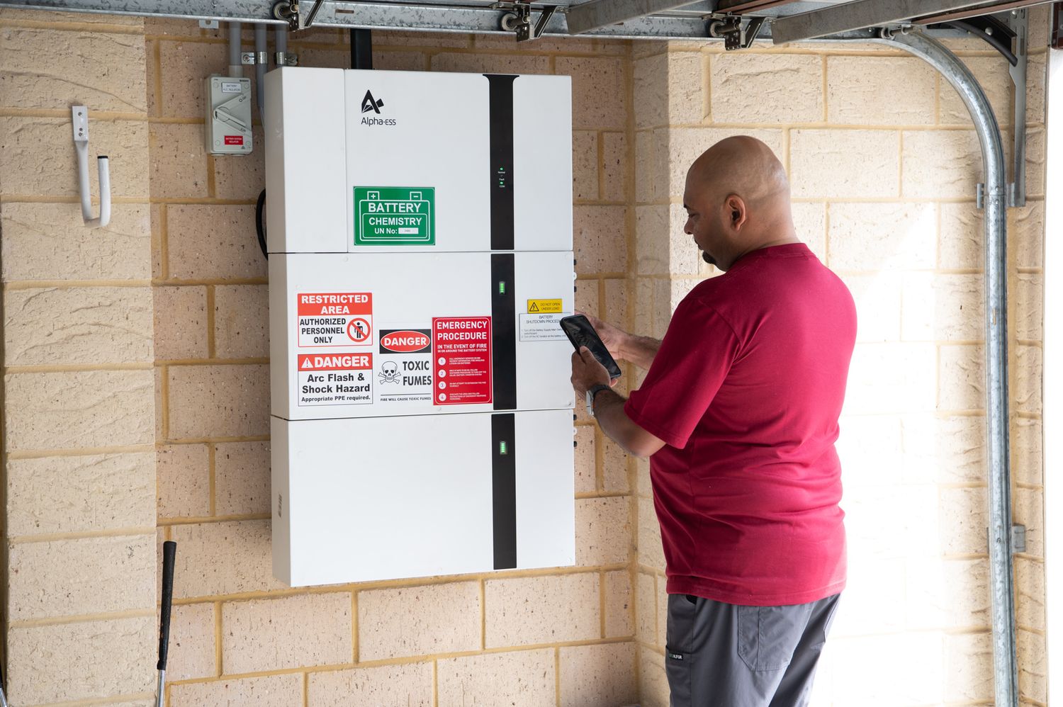 A person in a red shirt tests electrical equipment on a beige block wall mounted with industrial panels.