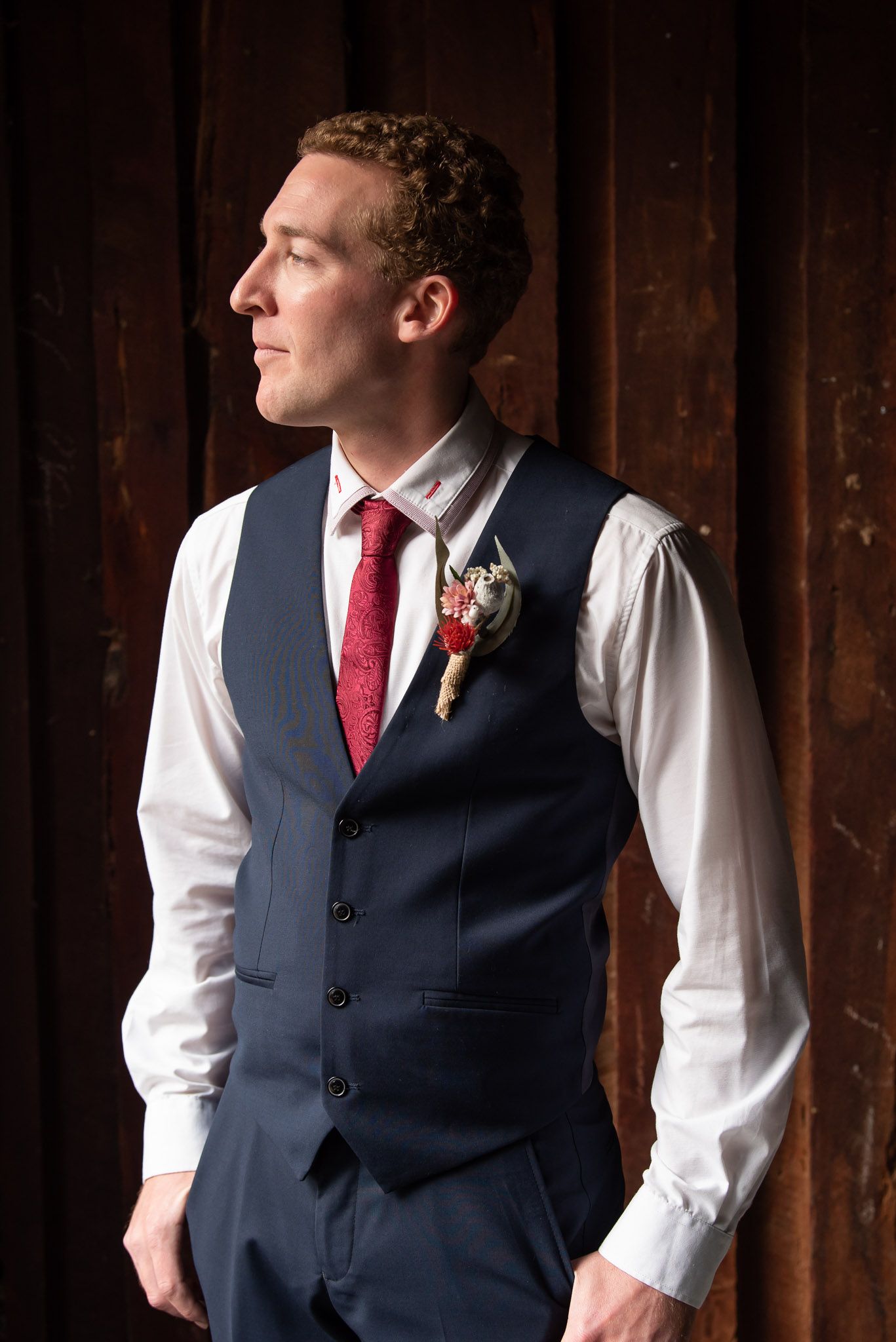 Close-up profile shot of a man wearing a navy vest and red tie against a dark wooden wall background.