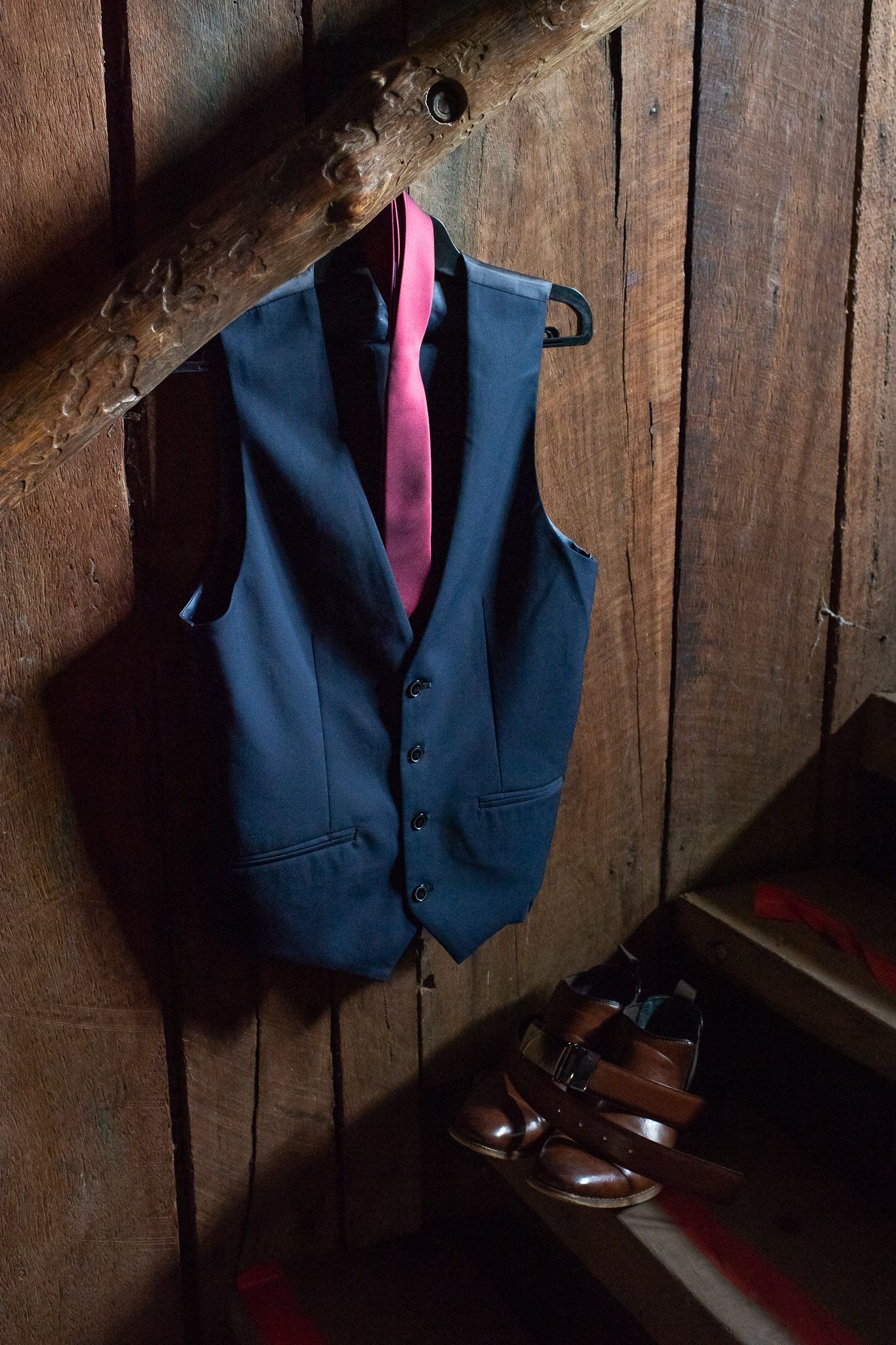 Navy blue suit vest with pink tie hanging on wood wall above brown leather dress shoes.