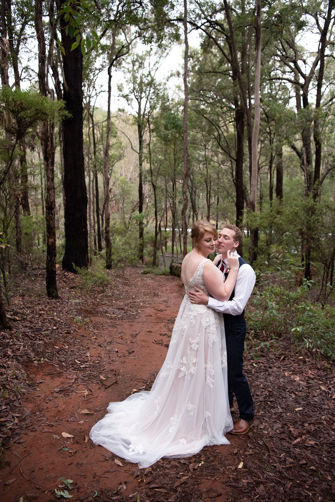 A romantic couple in wedding attire embrace on a winding dirt path through a serene eucalyptus forest.